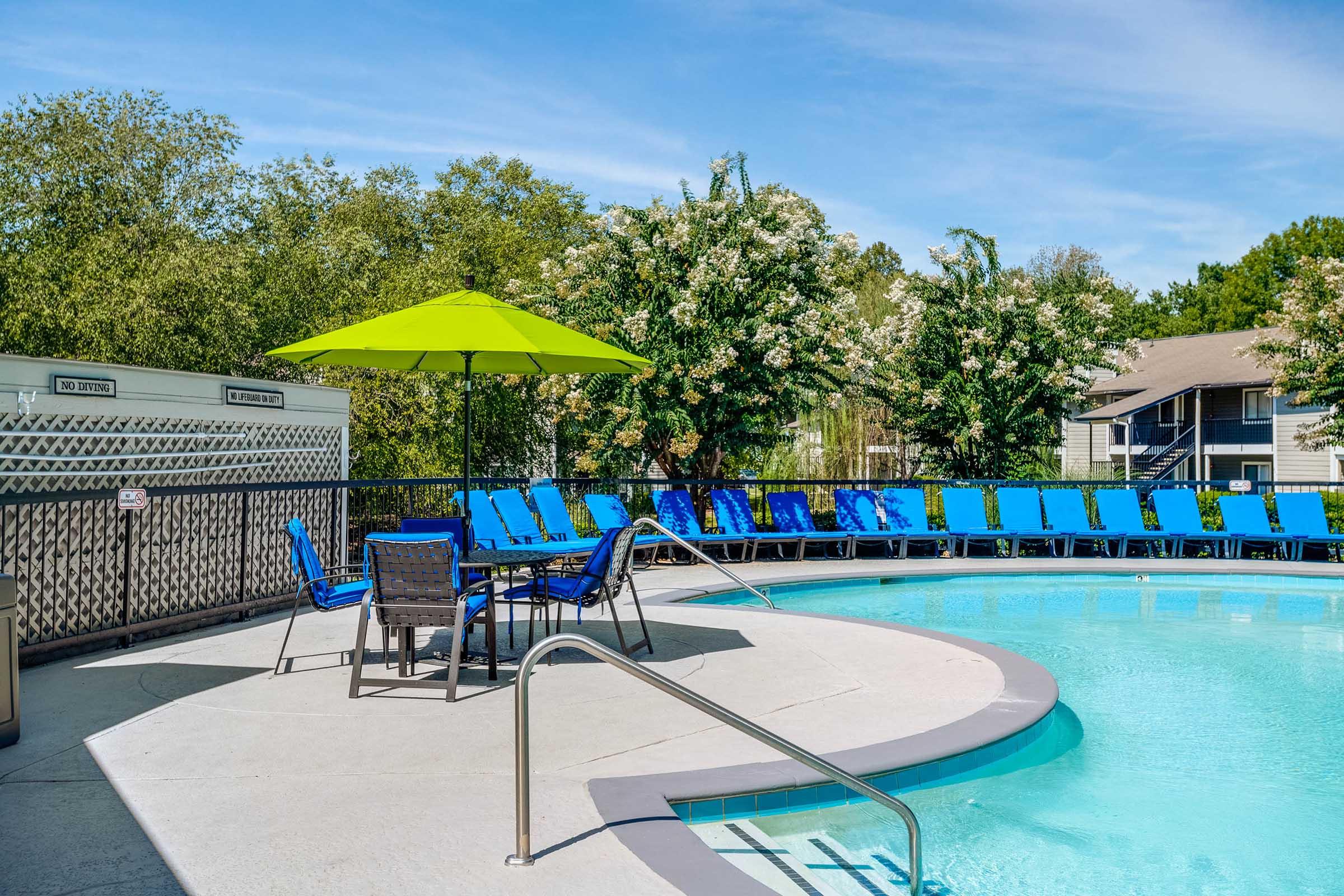 A bright outdoor pool area featuring a circular swimming pool, surrounded by blue lounge chairs. An umbrella in vibrant green provides shade over a table with chairs. Lush greenery and trees are visible in the background against a clear blue sky.
