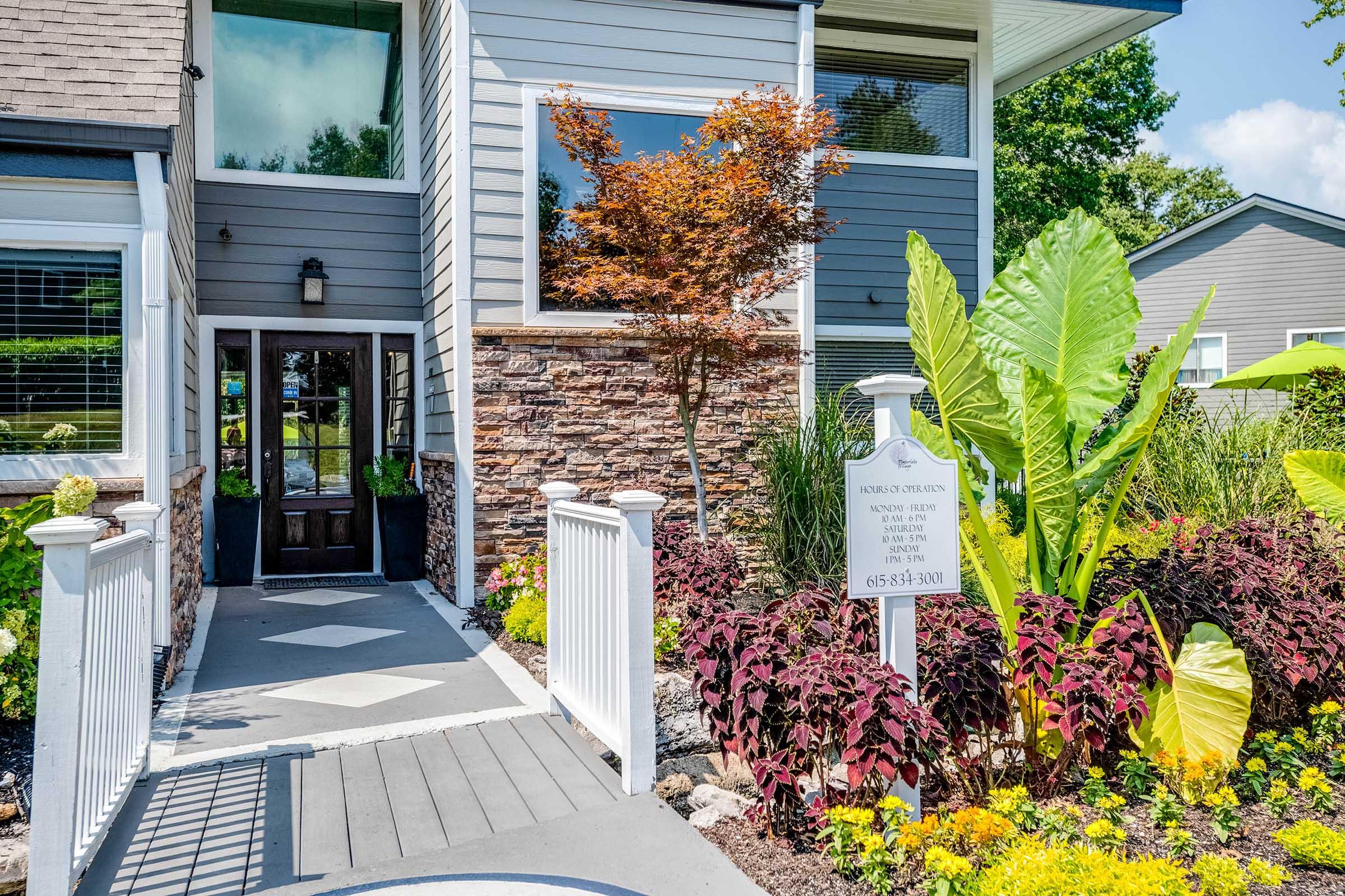A welcoming entrance of a modern building featuring a gray facade, a stone accent wall, and lush landscaping. The pathway is bordered by vibrant plants, including tropical foliage and colorful flowers, leading up to a dark front door. A sign is visible near the entrance with contact information.