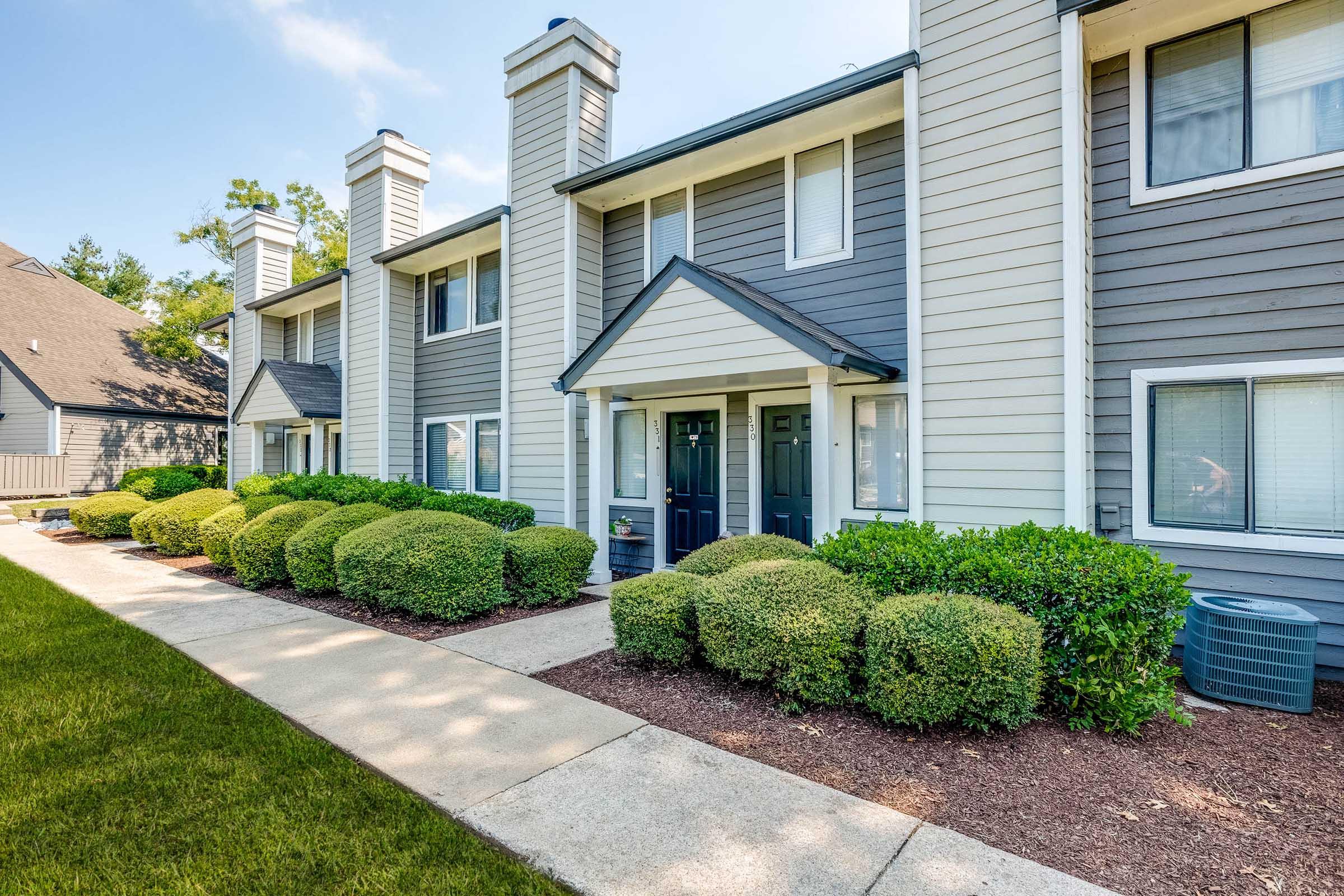 A row of modern townhouses with a well-maintained pathway and landscaping. The buildings feature a mix of light and dark siding, with neatly trimmed bushes in front. The sky is clear, and the area is surrounded by trees, creating a peaceful residential environment.