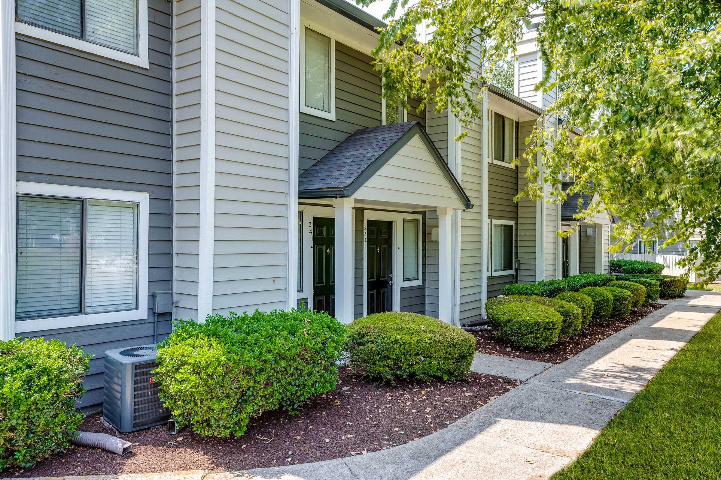 A pathway leading to a series of modern townhouses, featuring a well-manicured lawn with neatly trimmed shrubs on either side. The buildings have a mix of gray siding and white trim, with shaded entryways. Sunlight filters through the leafy trees nearby, creating a bright and inviting atmosphere.