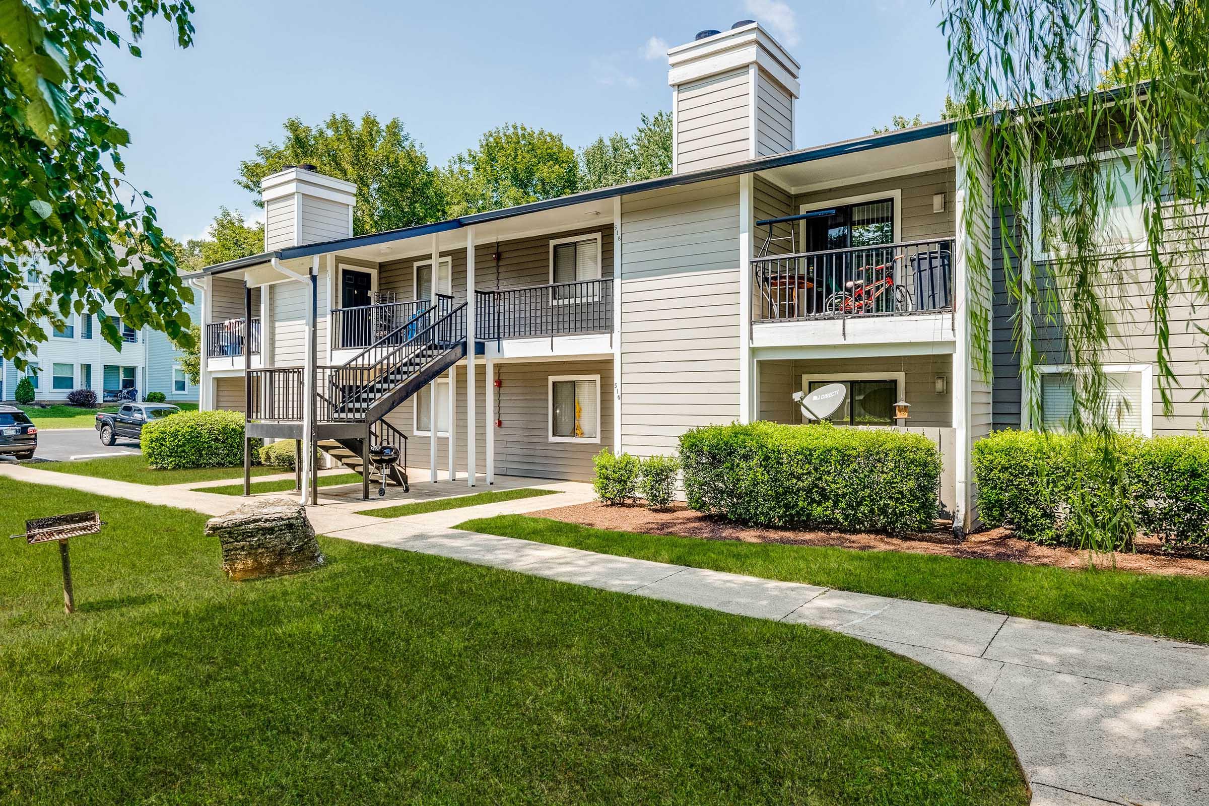 Two-story apartment building with a gray exterior, featuring balconies and black railings. Green lawn with neatly trimmed bushes in front. Sidewalk leading to the building and a signpost nearby. Clear blue sky and trees in the background.