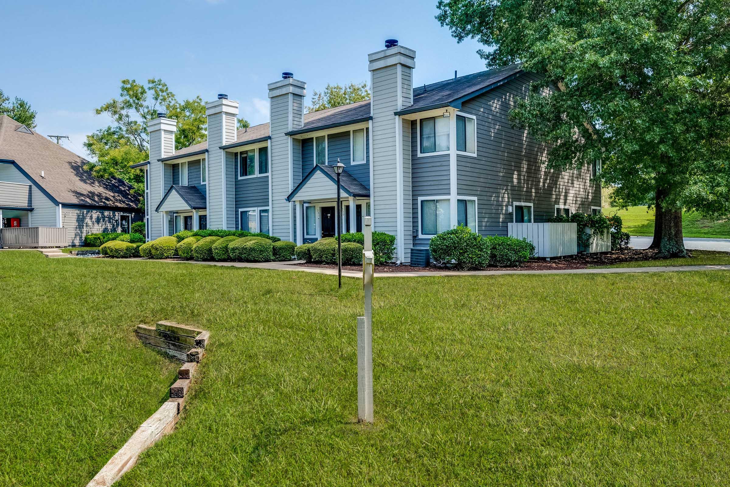 A modern apartment complex with gray siding and white trim, featuring three units, green landscaping, well-manicured lawns, and a clear blue sky overhead. A tall lamp post stands in the foreground, and decorative bushes line the path to the entrance.