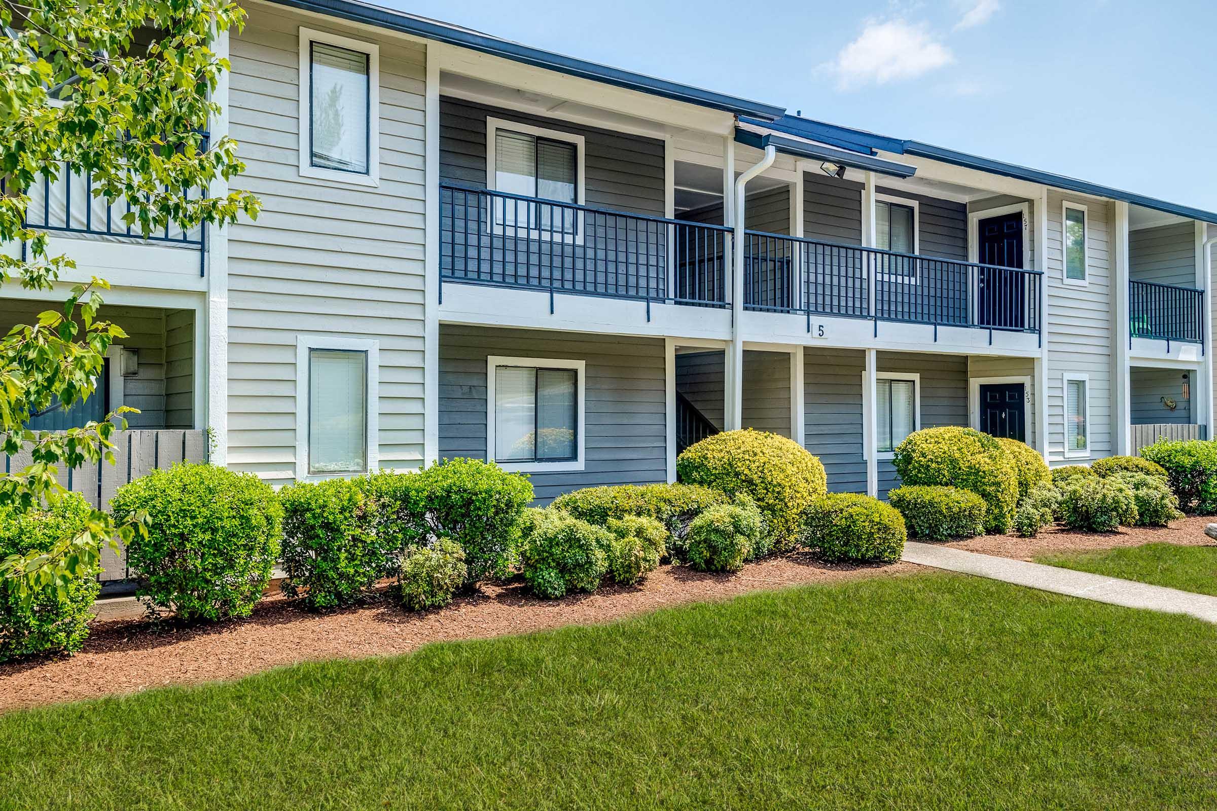 Two-story apartment building with gray siding and blue accents. The exterior features balconies, well-maintained shrubs, and a grassy area in front. Clear blue sky in the background enhances the pleasant setting. The pathway leads to the entrance, creating a welcoming atmosphere.