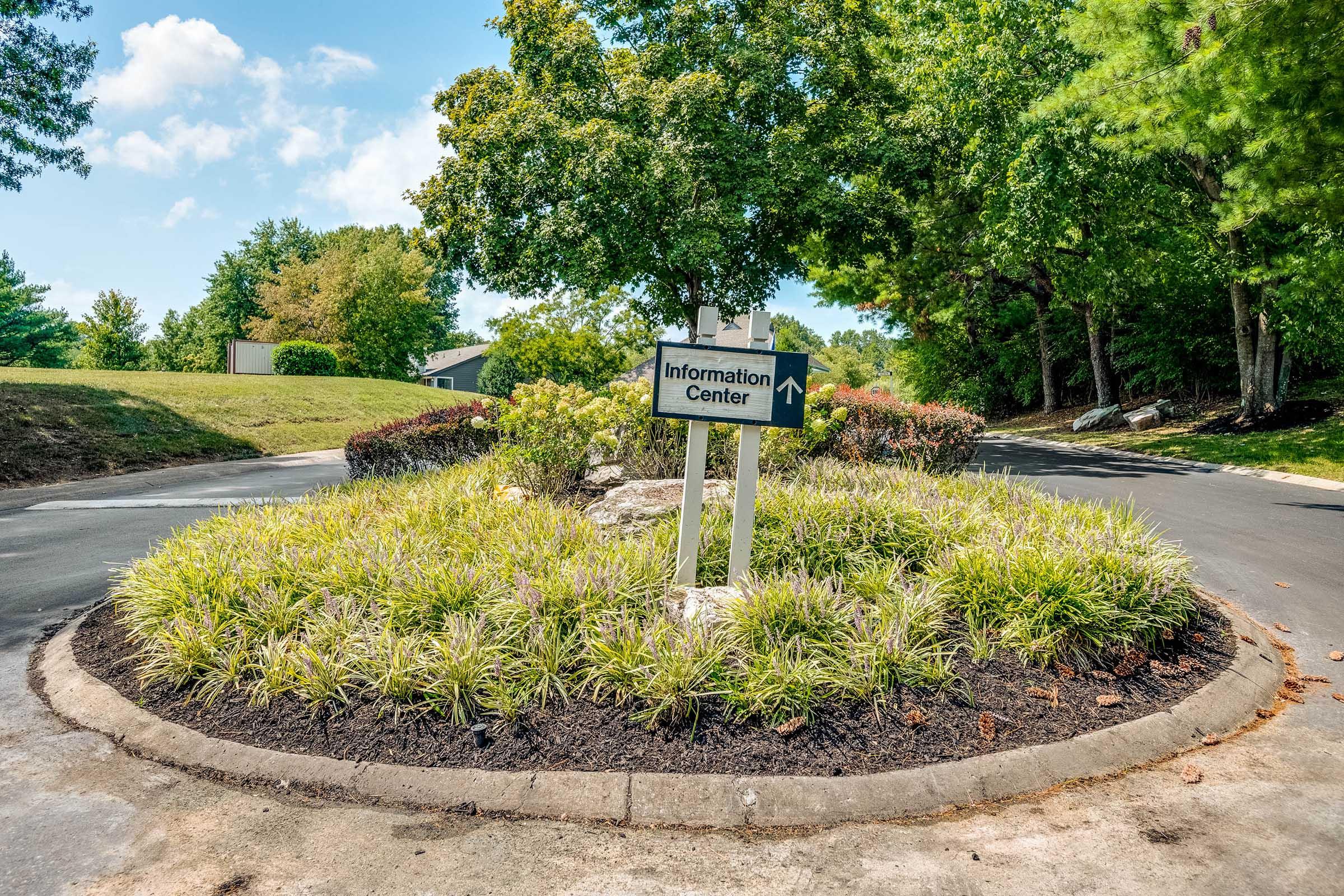 A signpost indicating "Information Center" with an arrow pointing right, surrounded by neatly landscaped greenery, including bushes and grass, in a sunny outdoor setting. The scene features trees in the background and a clear blue sky.