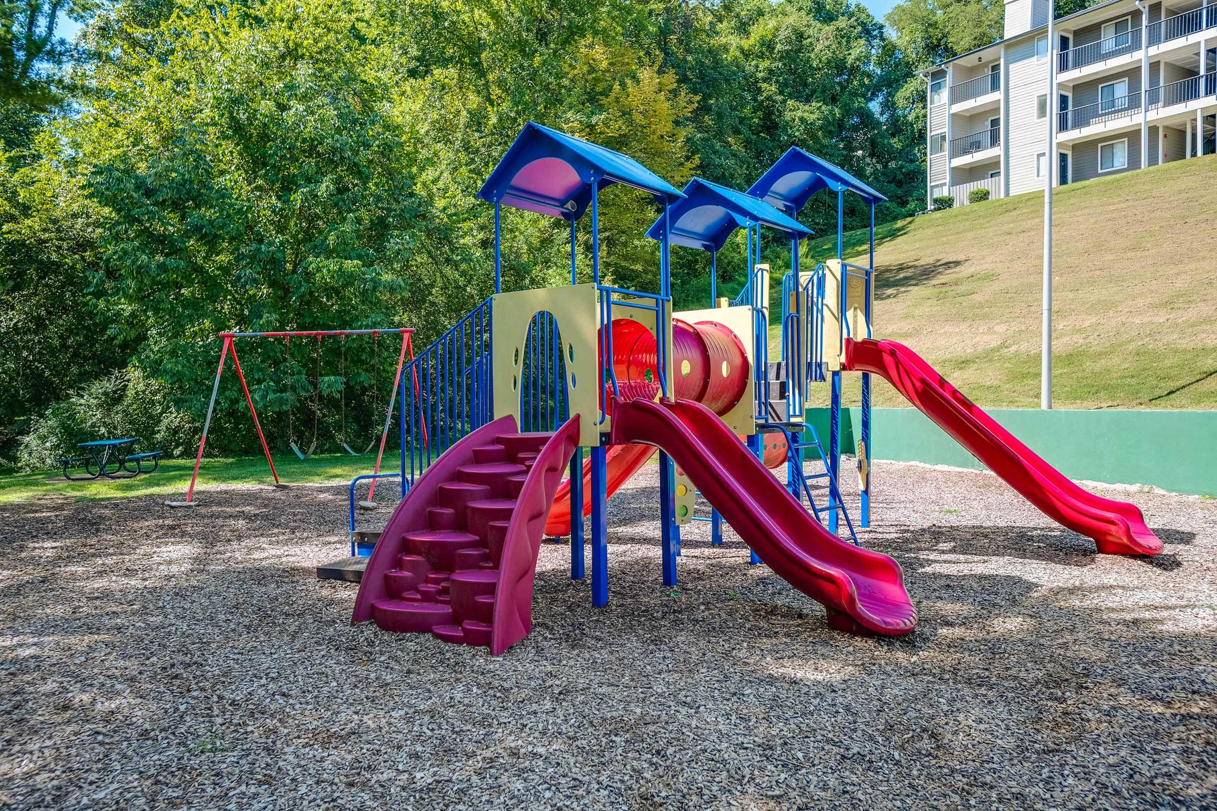 Colorful playground equipment featuring two red slides, climbing steps, and a series of overhead canopies. Surrounding the playground is a carpet of wood chips, with a swing set visible in the background and green grass on a slope. There are trees and a multi-story building in the distance.