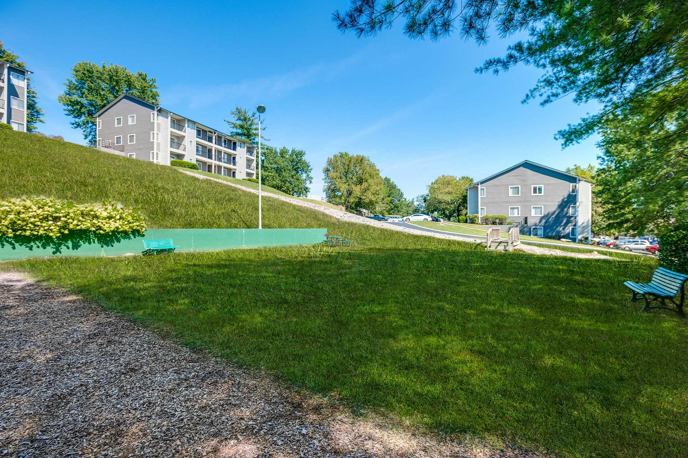 A grassy area with a gentle slope, featuring benches. In the background, there are two multi-story residential buildings surrounded by trees. The sky is clear and blue, indicating a sunny day.
