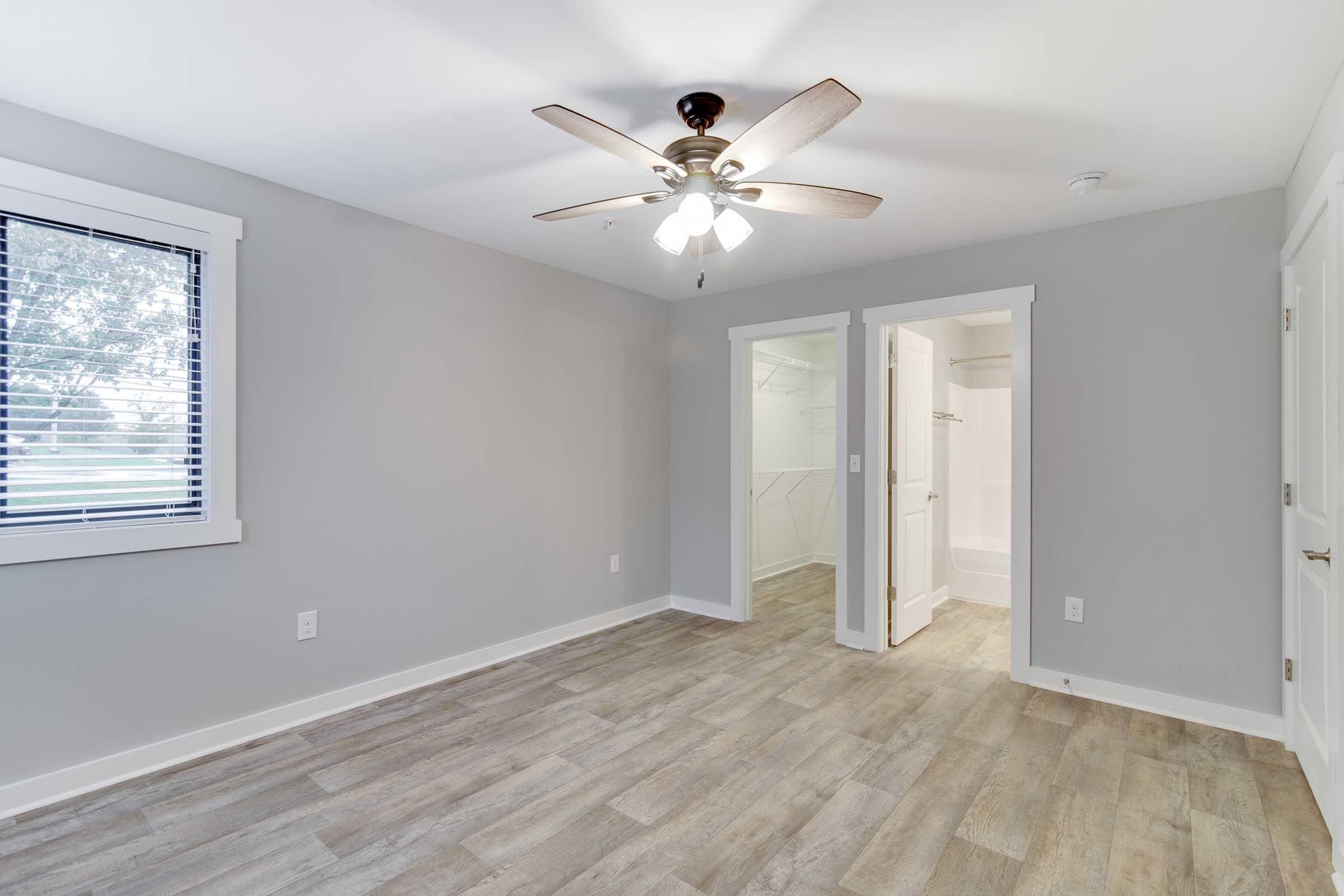 A modern interior of an empty room featuring light gray walls, a ceiling fan with wooden blades, and a large window with blinds. To the right, there are open doors leading to a bathroom and a closet. The floor is styled with light wooden planks, creating a spacious and inviting atmosphere.