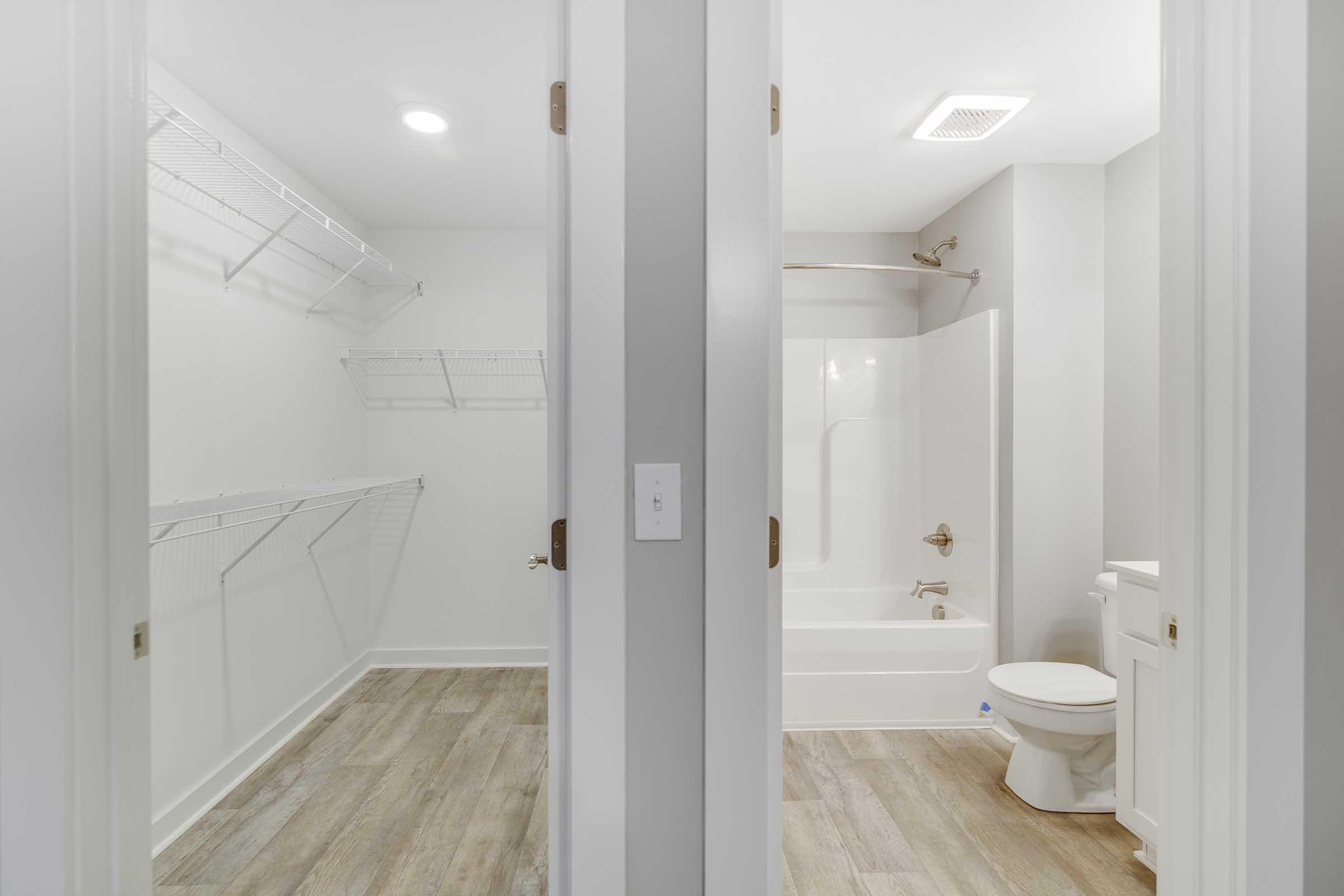 A modern bathroom with a bathtub and shower combo, next to a spacious walk-in closet featuring wire shelving. The walls are painted in a light gray color, and the flooring is a light, wood-like material. Soft white lighting illuminates the space.