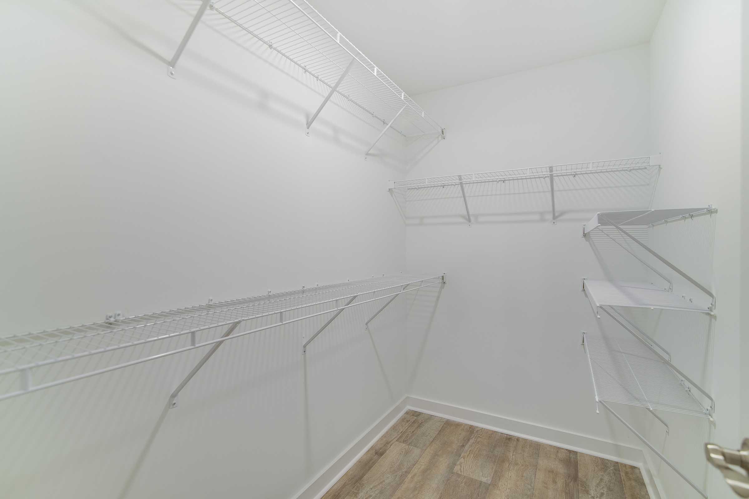 Empty walk-in closet featuring white wire shelving mounted on the walls. The room has a light-colored wall and wooden flooring, creating a clean and spacious appearance.