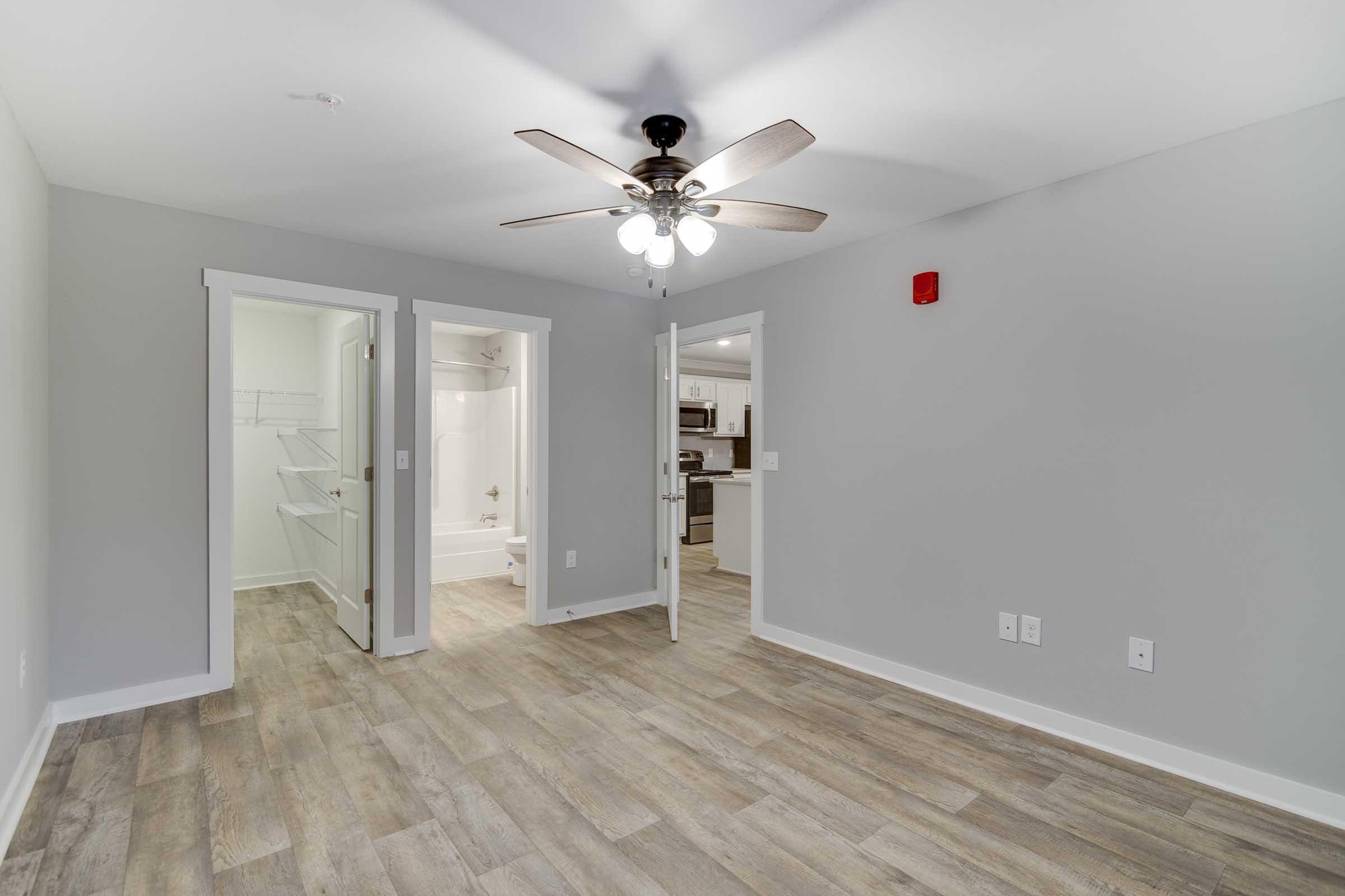 A spacious interior of an apartment featuring a ceiling fan, light-colored walls, and wood-like flooring. Two open doors lead to a bathroom and a kitchen area. The room is well-lit and modern, creating a welcoming atmosphere.