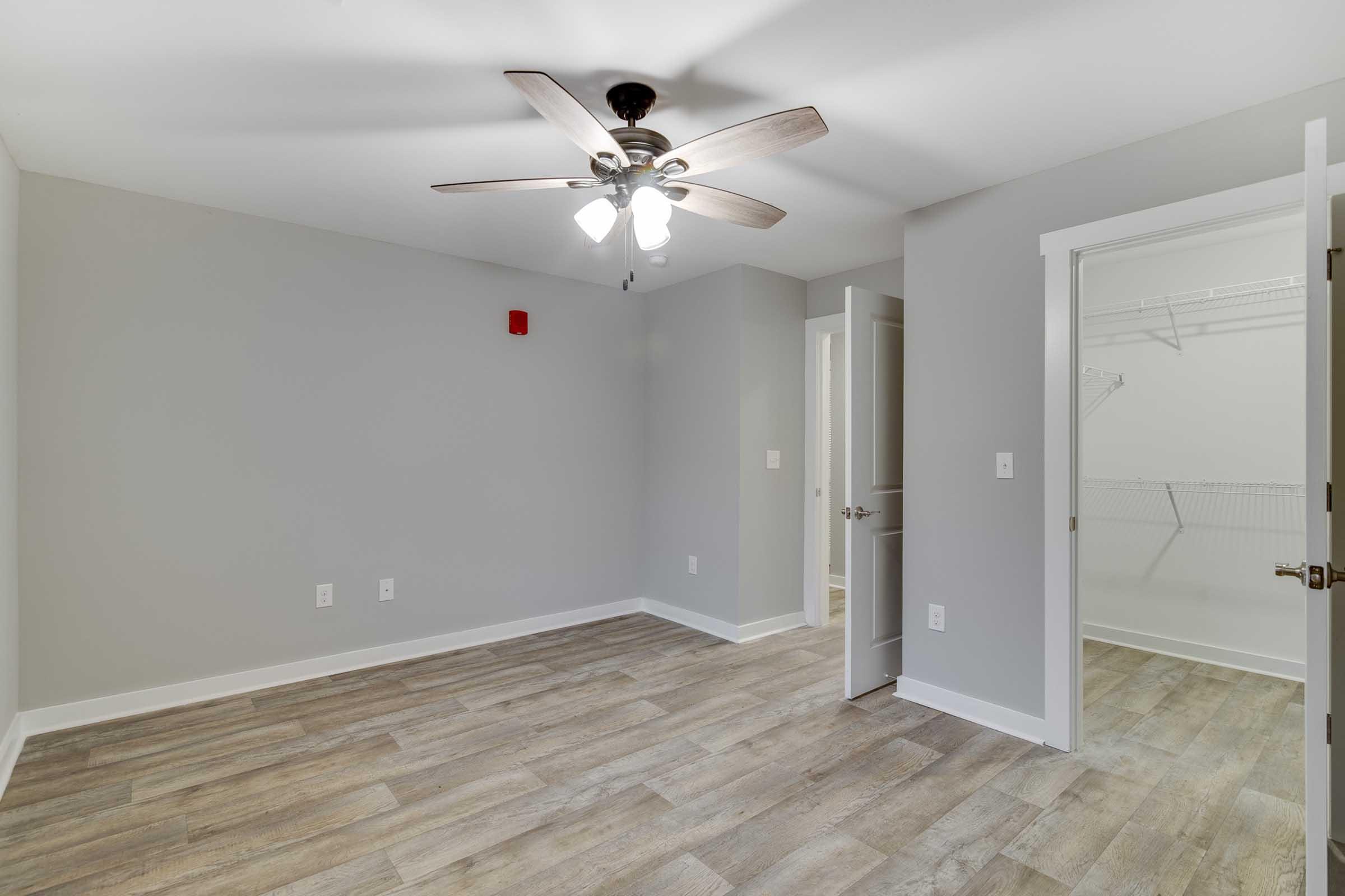 A neutral-colored bedroom featuring a ceiling fan with light fixtures, a closet with a wire shelf, and light-colored wooden flooring. The walls are painted in a light gray tone, and there is an open door leading to another room or hallway, providing a bright and spacious feel.