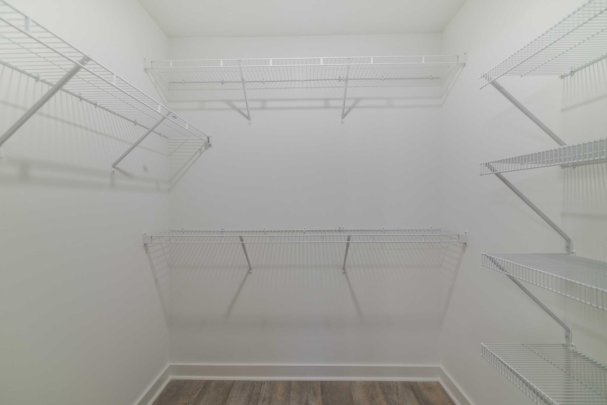 Empty walk-in closet with white wire shelving against plain white walls. Flooring is light brown wood-like material. The space is well-lit and uncluttered, providing a blank canvas for storage or organization.