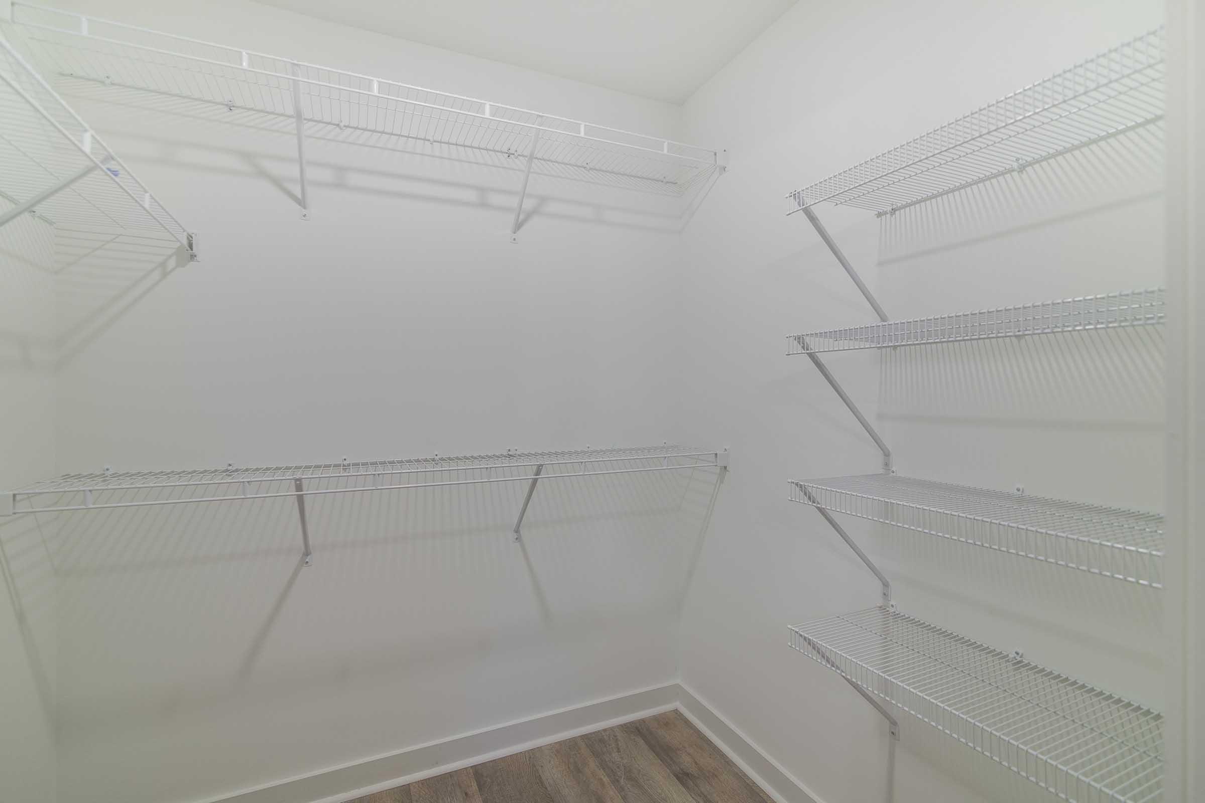 A well-lit empty closet with white walls and several metal wire shelving units on three walls. The floor has a wooden texture, and the shelves are arranged at different heights, creating an organized and spacious look. The overall atmosphere is clean and minimalistic.