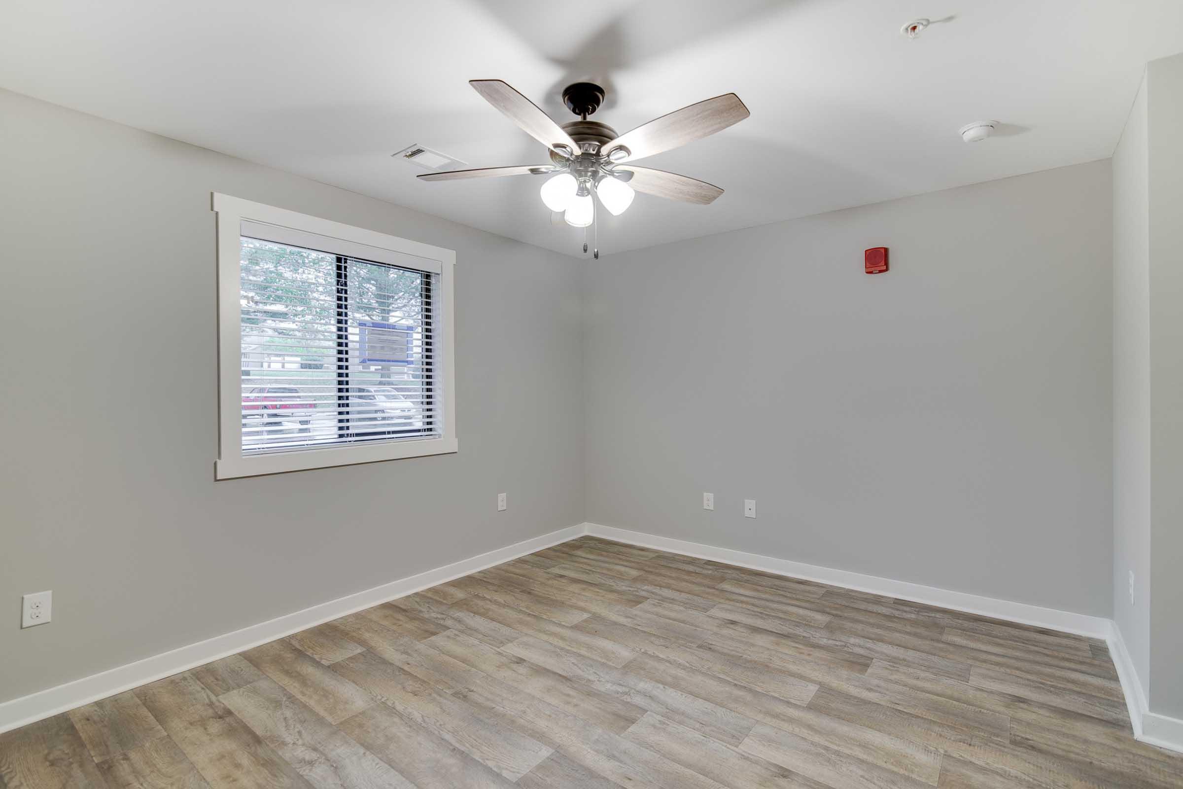 A vacant room with light gray walls, a ceiling fan with wooden blades, and a window with horizontal blinds. The flooring is a light wood look. There is a smoke detector on the ceiling and a fire alarm on the wall. The overall ambiance is bright and minimalistic.