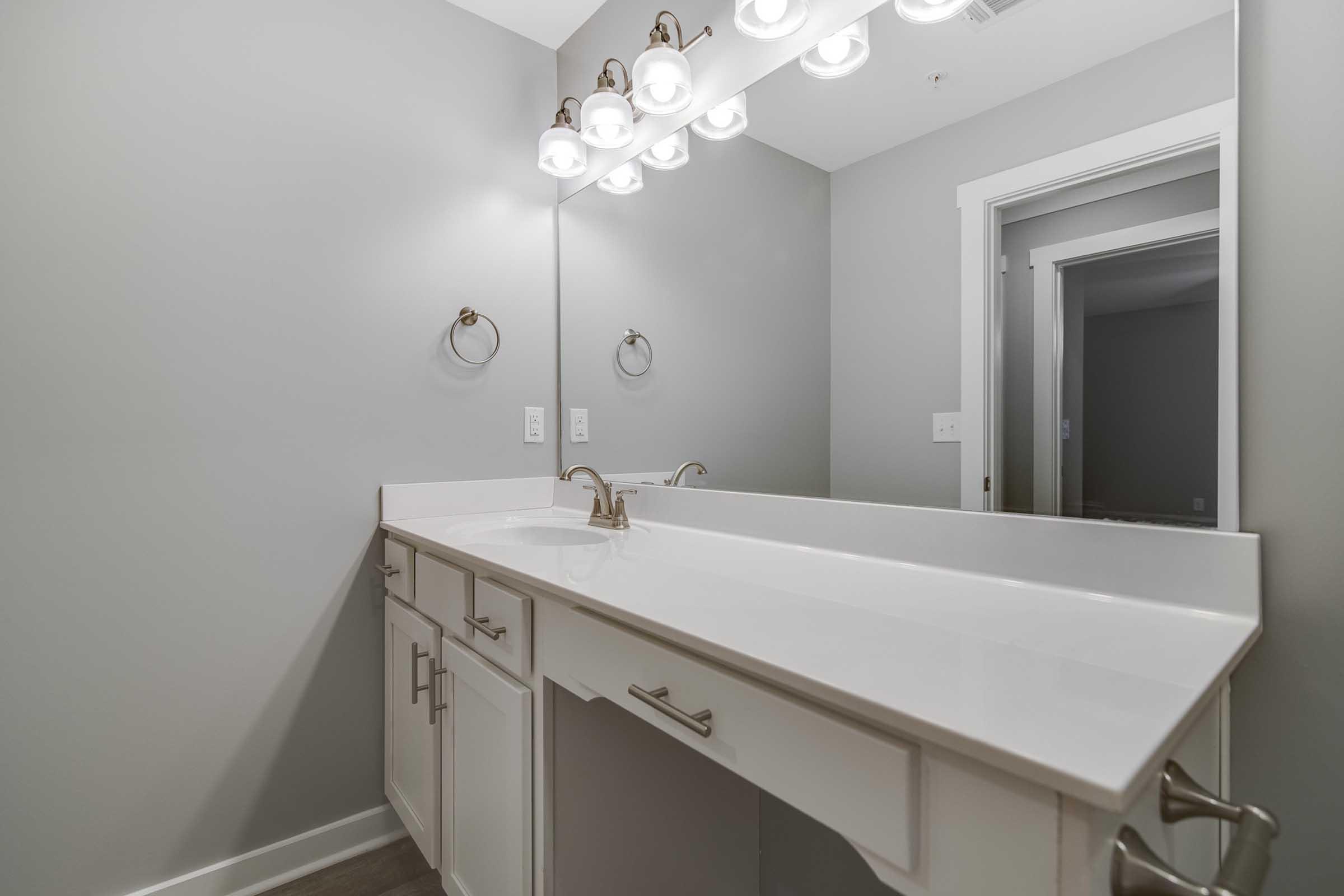 A modern bathroom with a white countertop and double sink, featuring a light fixture with five bulbs above the mirror. The walls are painted gray, and there are two towel rings on either side of the sink. A doorway is visible in the background, leading to another room.