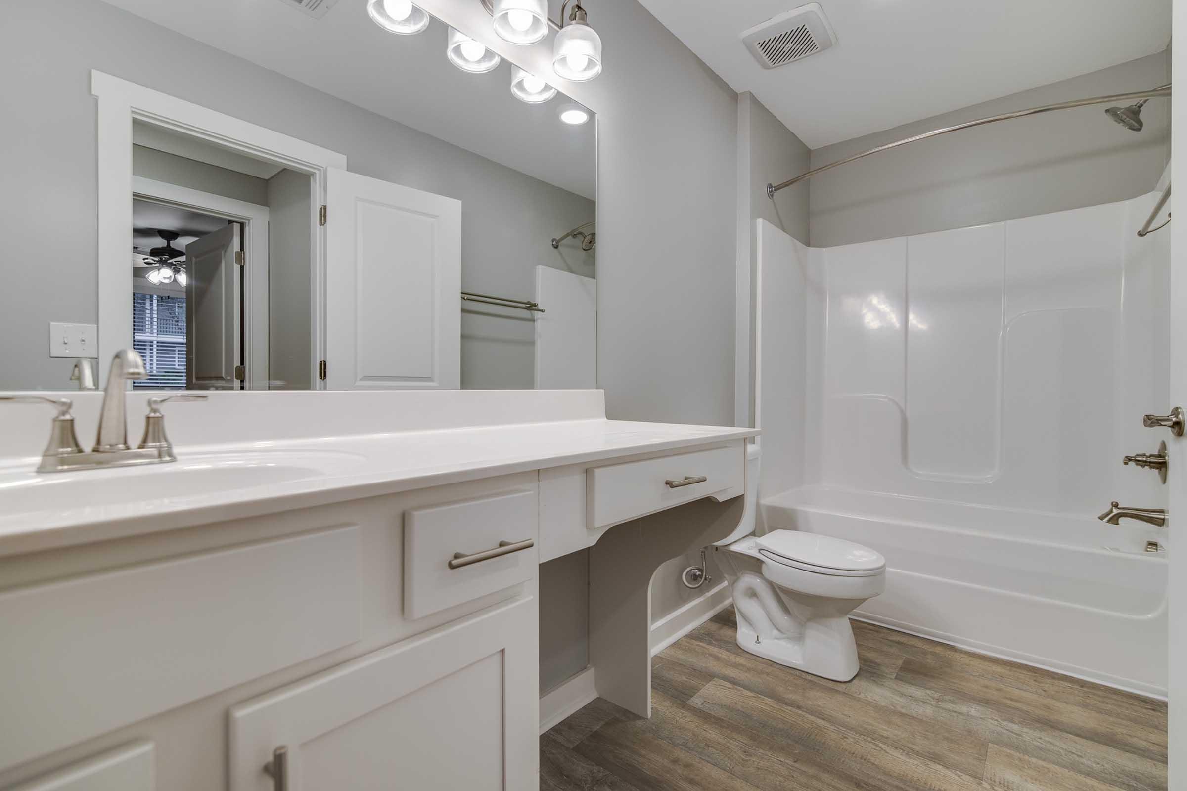 A modern bathroom featuring a white vanity with dual sinks, a large mirror, and multiple light fixtures. There is a white bathtub with a shower, a toilet, and light-colored wood flooring. The walls are painted in a neutral tone, and a door leads to another room in the background.
