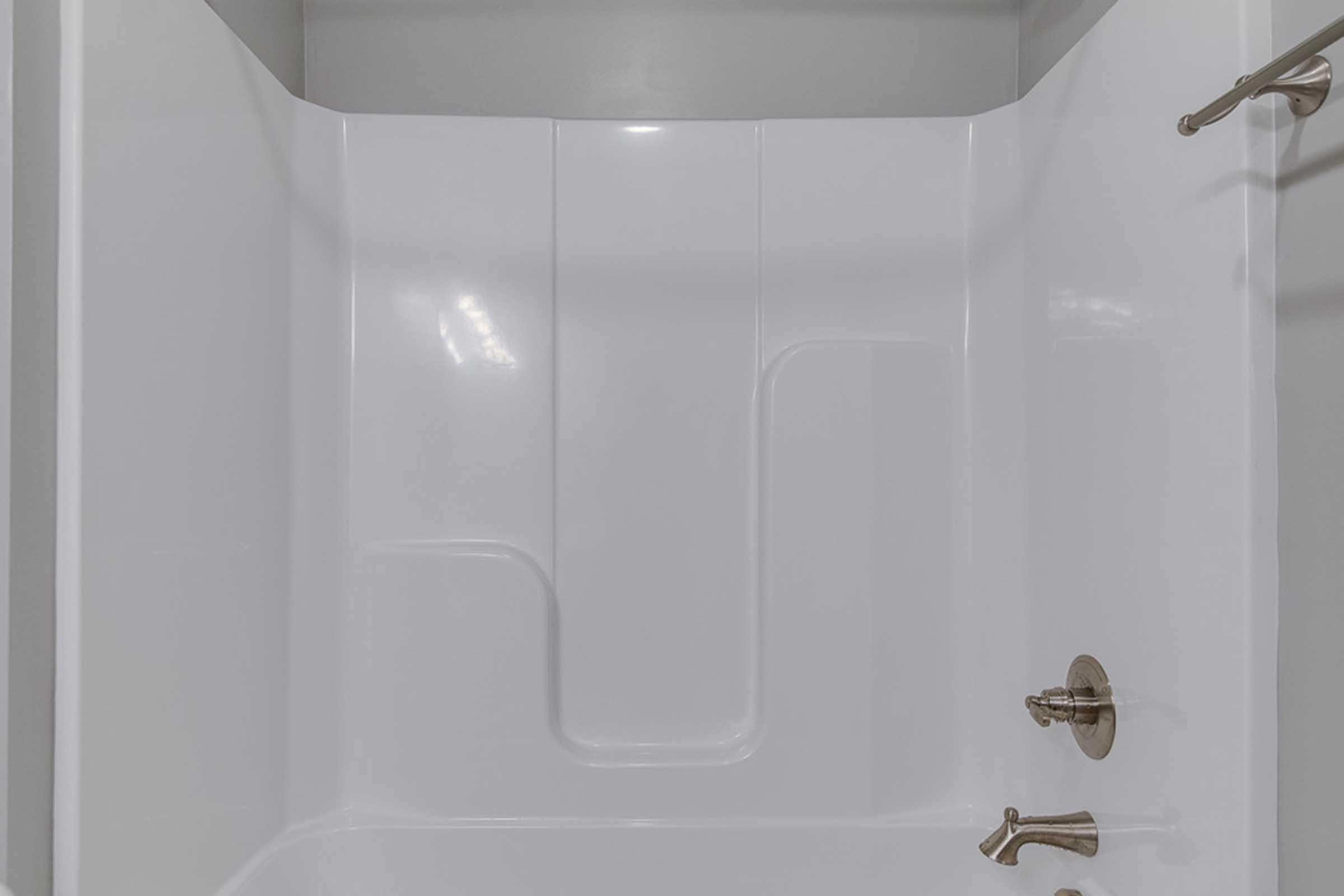 A clean, modern shower stall with a smooth white surface, featuring a built-in seat. The walls are curved, and there are two brass fixtures: one for controlling water temperature and the other for the showerhead. The overall aesthetic is minimalistic and well-maintained.