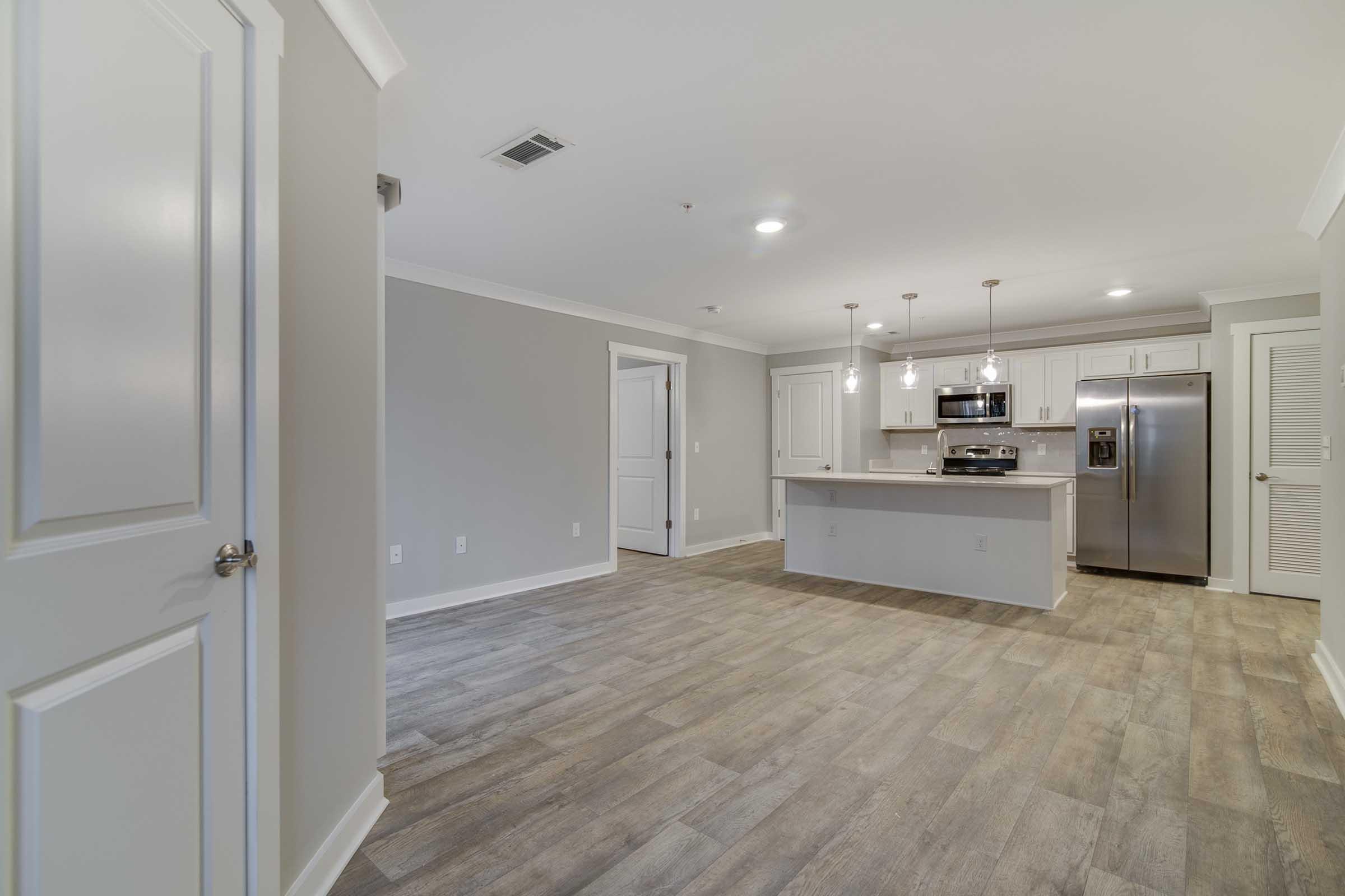 Modern open-concept kitchen and living area featuring light gray walls, recessed lighting, and wood-look tile flooring. The kitchen includes white cabinetry, stainless steel appliances, and a central island with pendant lighting. Adjacent is a door leading to a storage area.