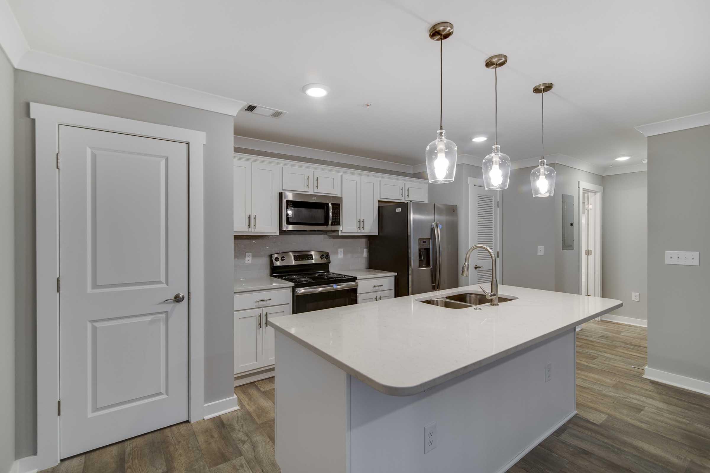 Modern kitchen featuring white cabinetry, stainless steel appliances, and a large island with a sink. Three pendant lights hang above the island, providing ample lighting. The flooring is a warm wood-tone, and the walls are painted in a soft gray, creating a bright and inviting atmosphere. A door leads to another room.