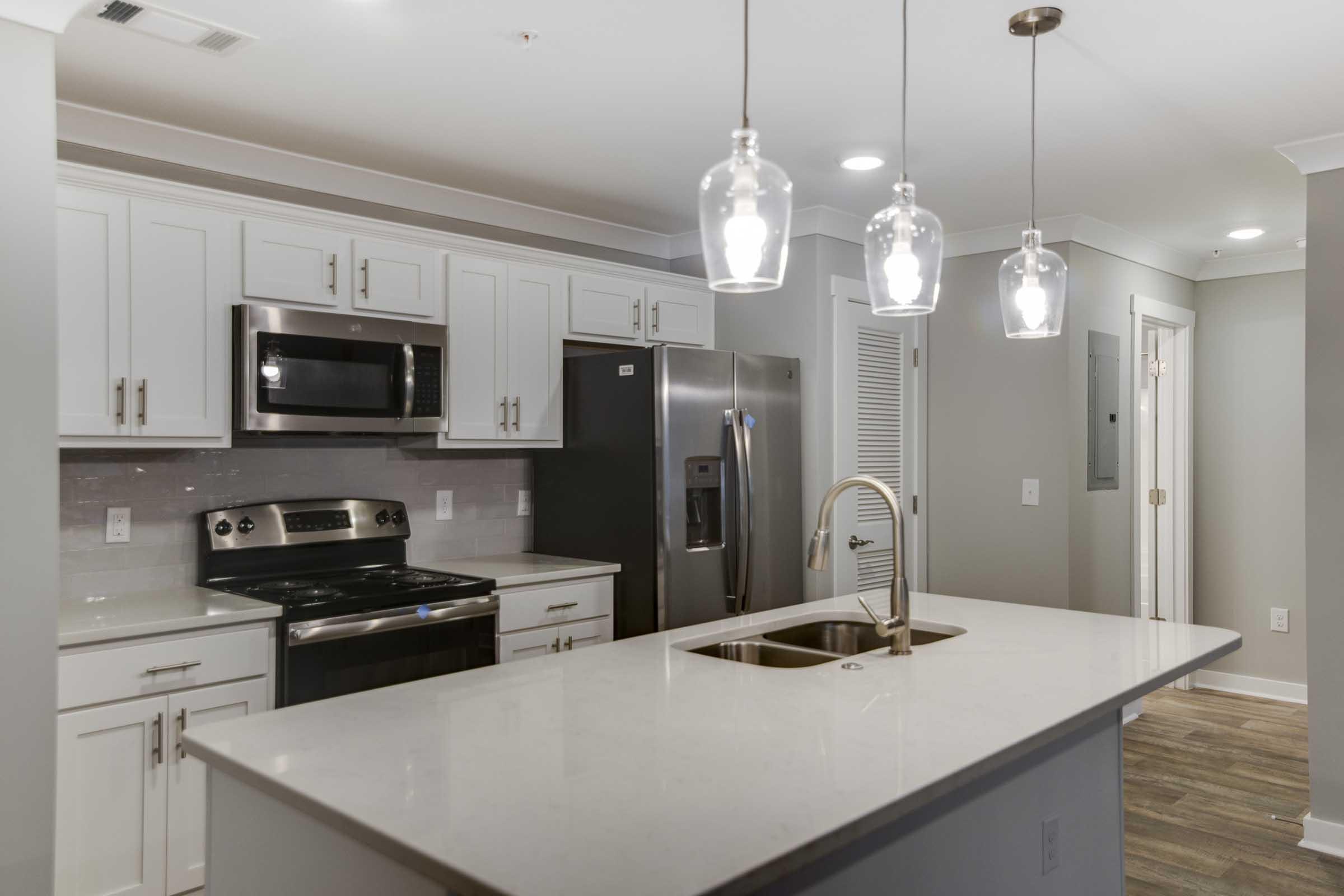 Modern kitchen featuring white cabinetry, stainless steel appliances including a stove and refrigerator, and a large island with a sink. Pendant lights hang above the island, and the walls are painted in a soft gray tone, creating a stylish and inviting atmosphere.