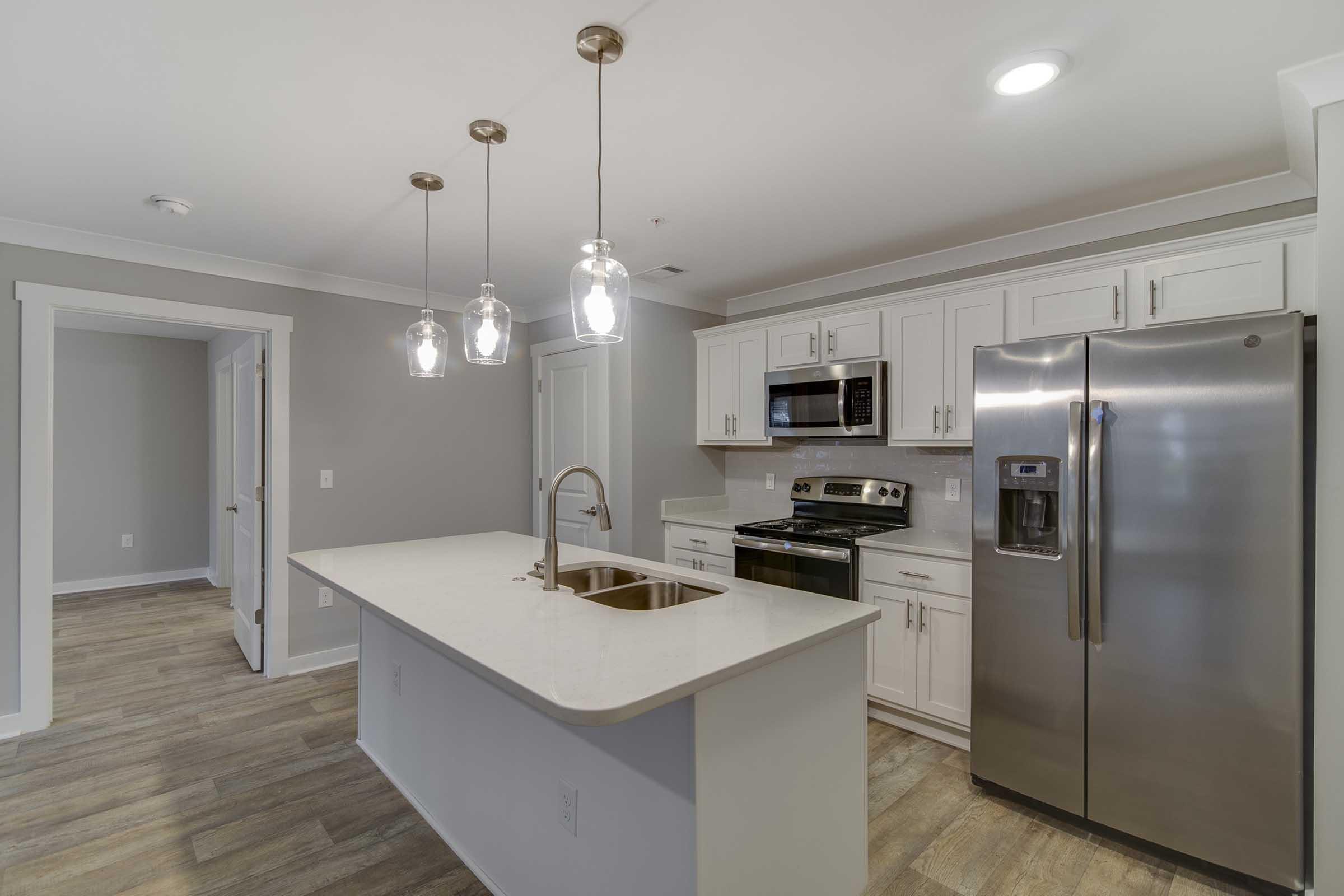 Modern kitchen featuring white cabinets, stainless steel appliances including a refrigerator and microwave, a large island with a sink, and pendant lighting above. The space is well-lit with natural light, and the flooring is a light wood finish. A doorway leads to another room in the background.