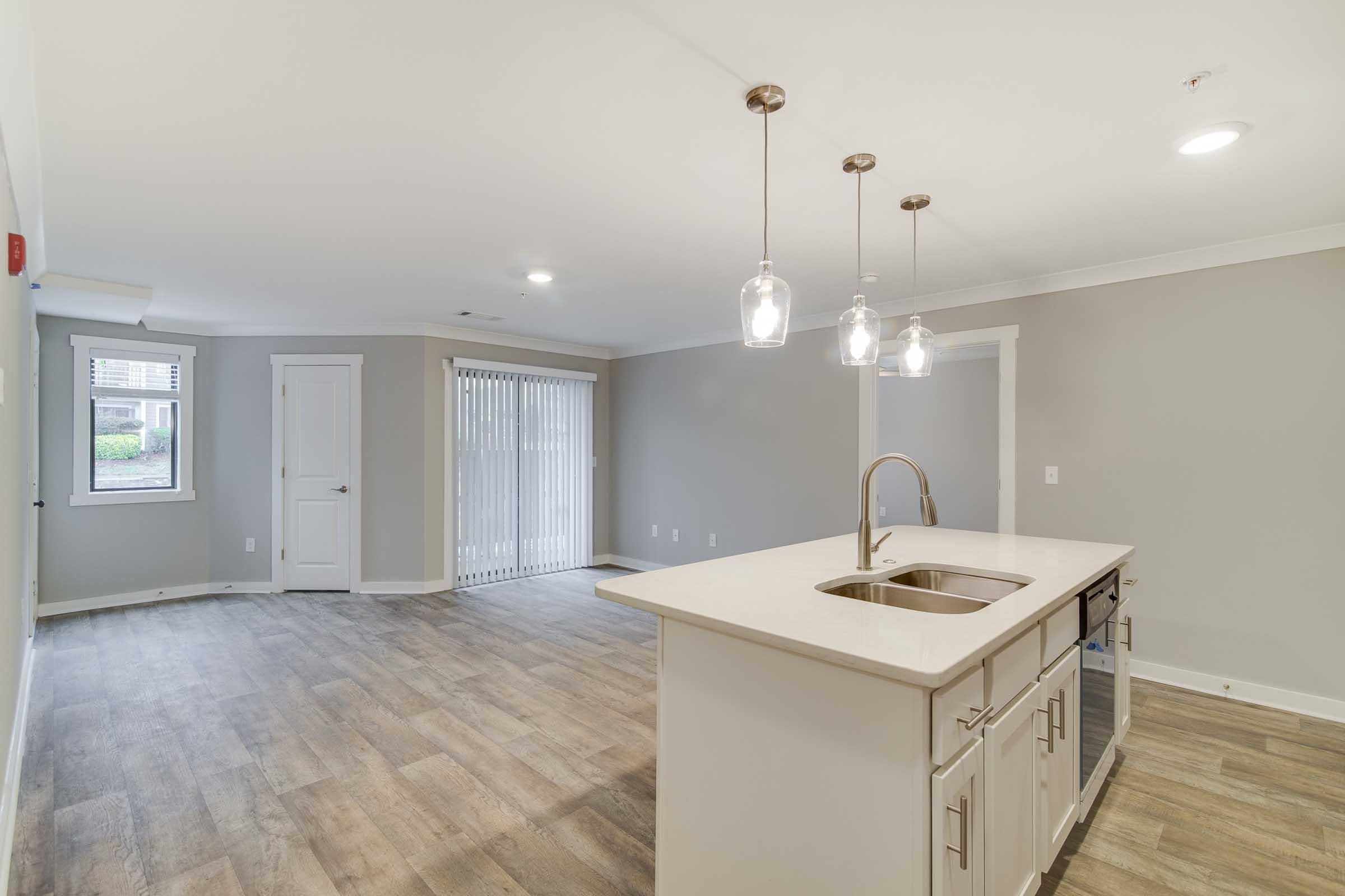 Spacious modern kitchen with a central island featuring a sink, surrounded by elegant pendant lights. Open living area with large windows and sliding glass doors, leading to a patio. Neutral wall colors and wood-like flooring create a bright and inviting atmosphere.