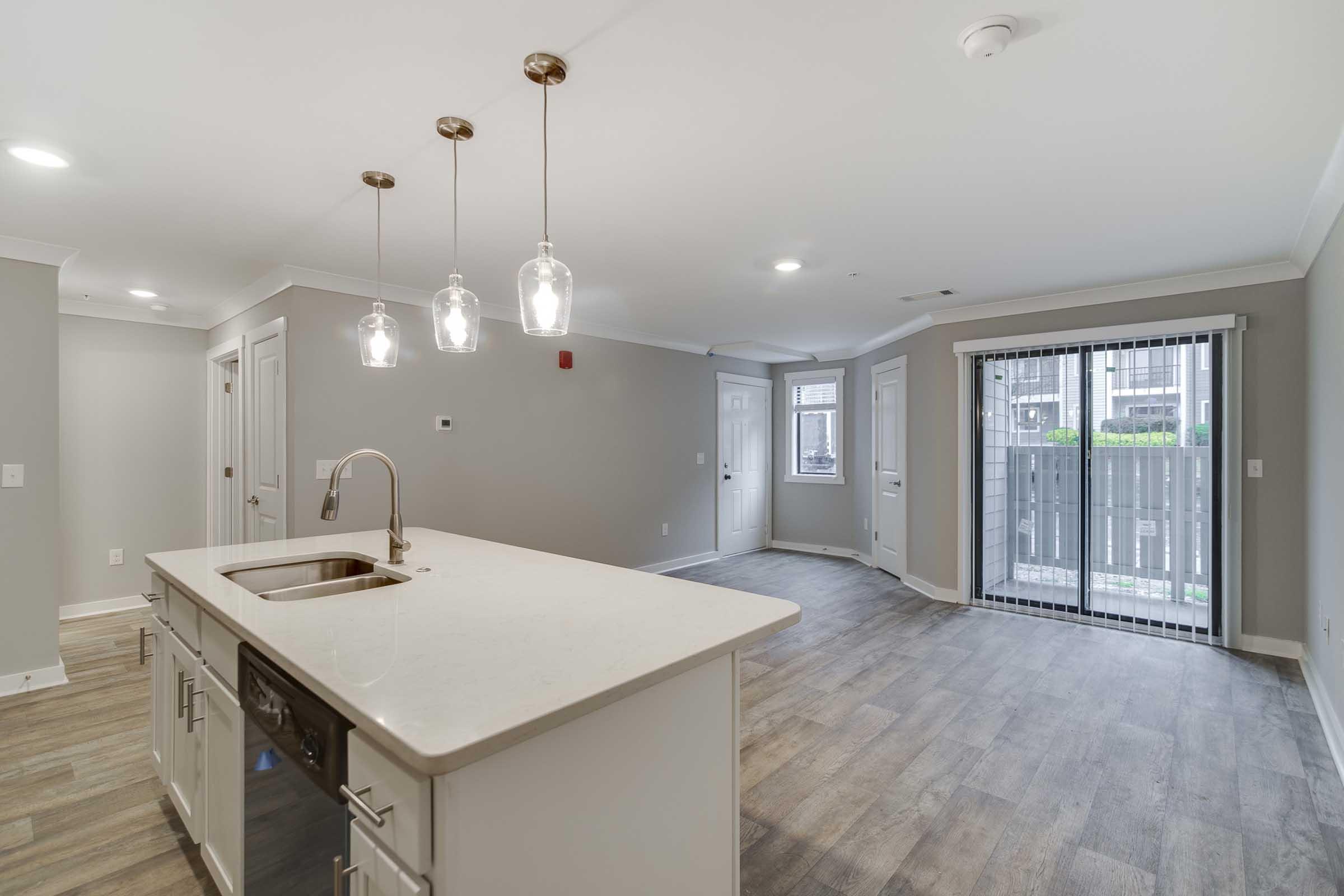 Modern interior of a spacious apartment featuring an open-concept layout. A kitchen island with a sink and pendant lighting is in the foreground, while the living area has large windows and access to a balcony. Neutral wall colors and hardwood flooring create a bright, inviting atmosphere.