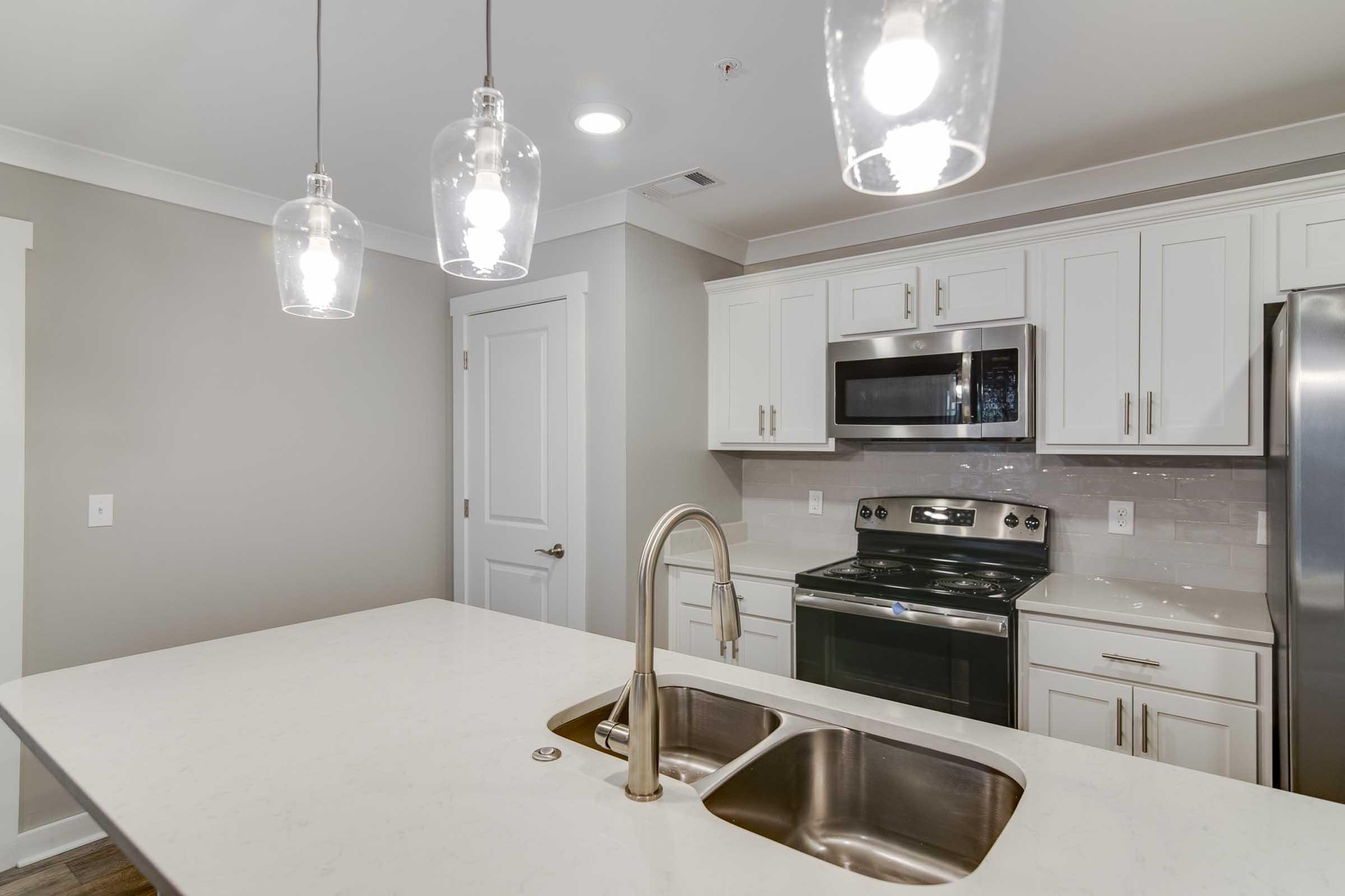 A modern kitchen featuring white cabinets, a stainless steel fridge, and an oven with a microwave above. The countertop is a light-colored marble, and there are two pendant lights hanging above the sink. The space is well-lit and has a minimalist design with a neutral color palette.