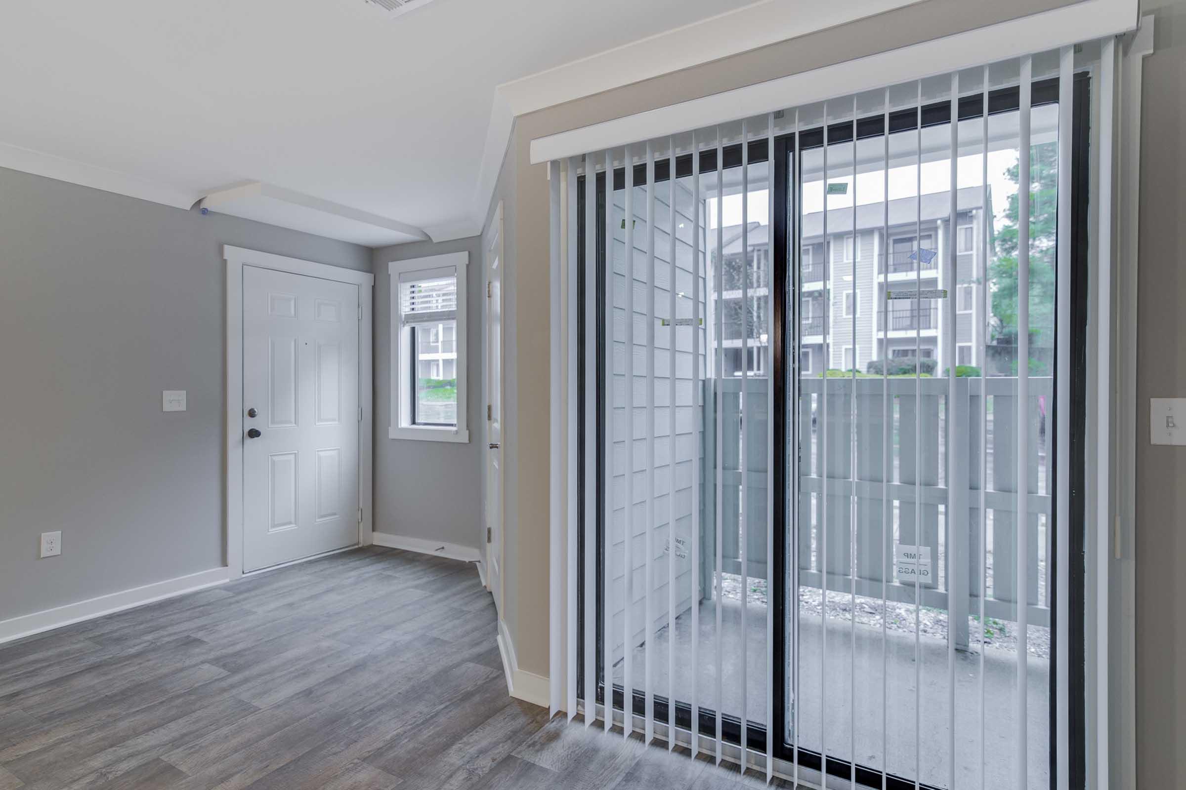 Interior view of a modern apartment featuring a door leading outside, a large sliding glass door with vertical blinds, and windows allowing natural light. The walls are painted in neutral tones, and there is a hardwood floor. The outside area appears to have a balcony or patio space.