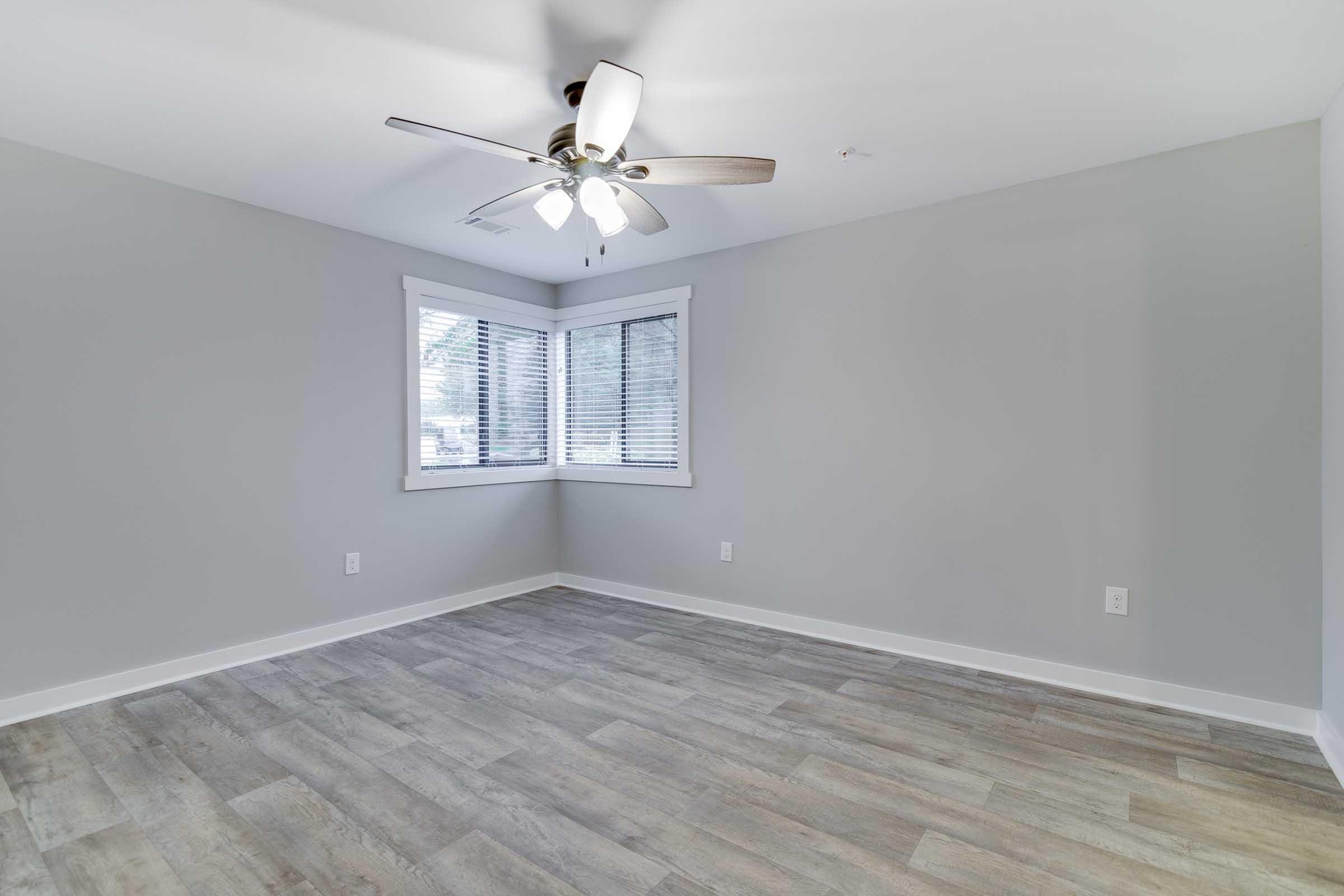 A spacious, empty room with light gray walls and a wooden floor. There is a ceiling fan with lights and a large bay window dressed with white blinds, allowing natural light to illuminate the space. The room has a clean and modern aesthetic, suitable for various furnishing options.