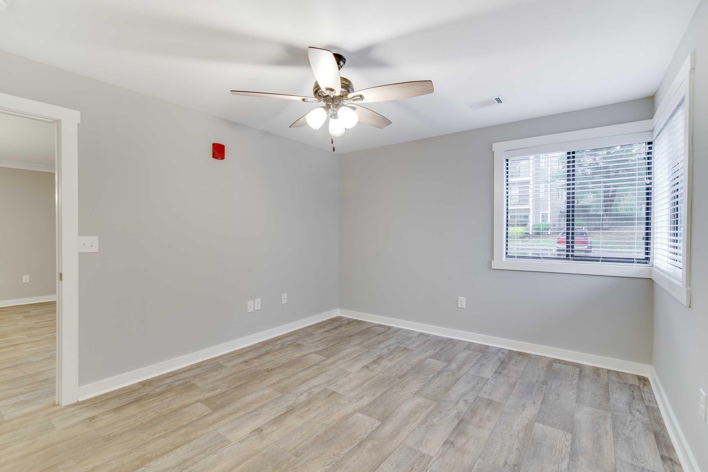 A bright, empty room featuring a ceiling fan with light fixtures, light gray walls, and laminate flooring. There is a window with blinds allowing natural light, and a small red alarm or light fixture on one wall. The space is clean and uncluttered, ideal for various uses.