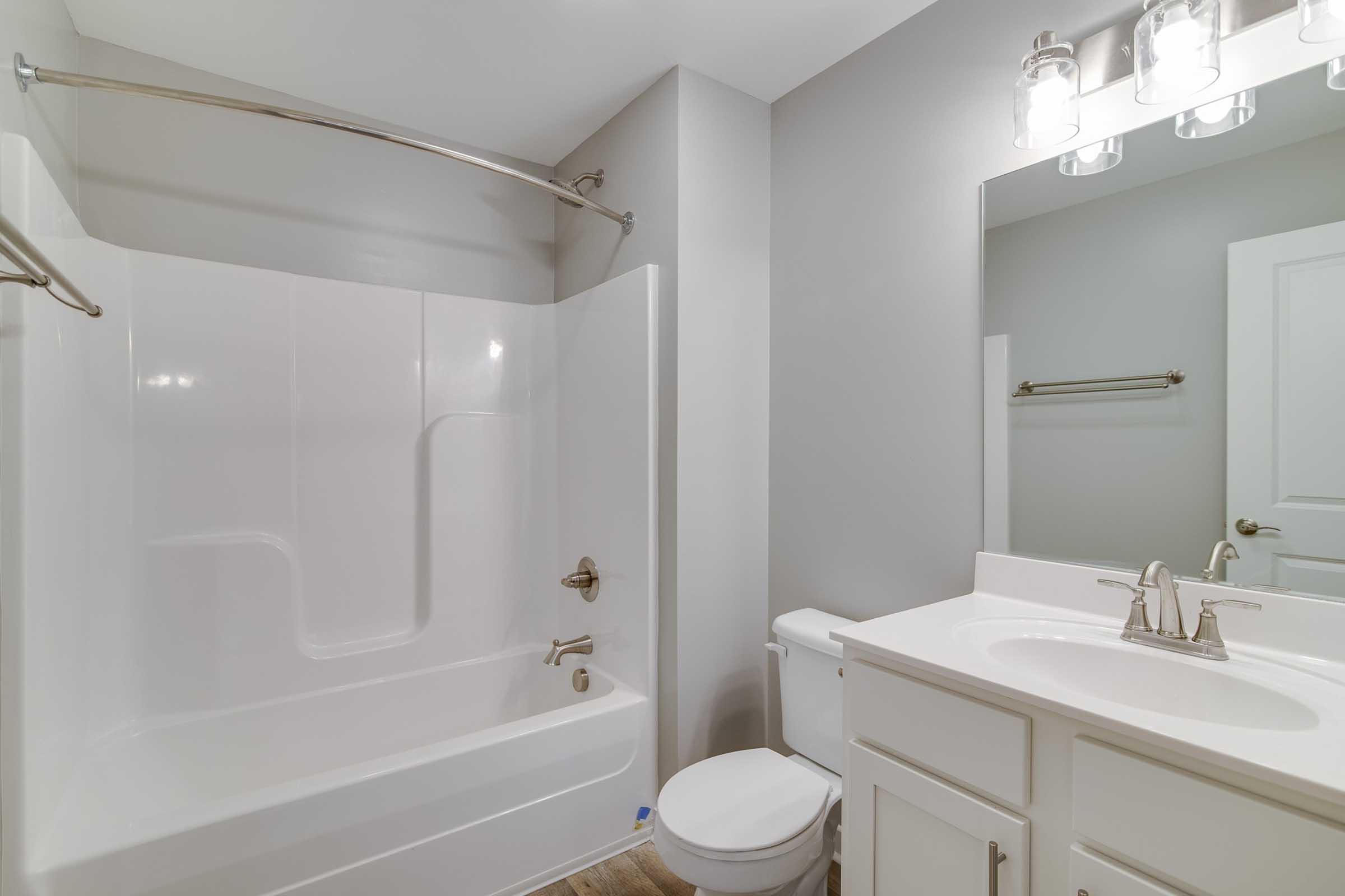 A clean, modern bathroom featuring a white bathtub with a shower rod, a matching toilet, and a stylish vanity with a sink. The walls are painted light gray, and there's a vanity mirror above the sink with bright light fixtures. The floor has a wood-like appearance, enhancing the overall contemporary design.