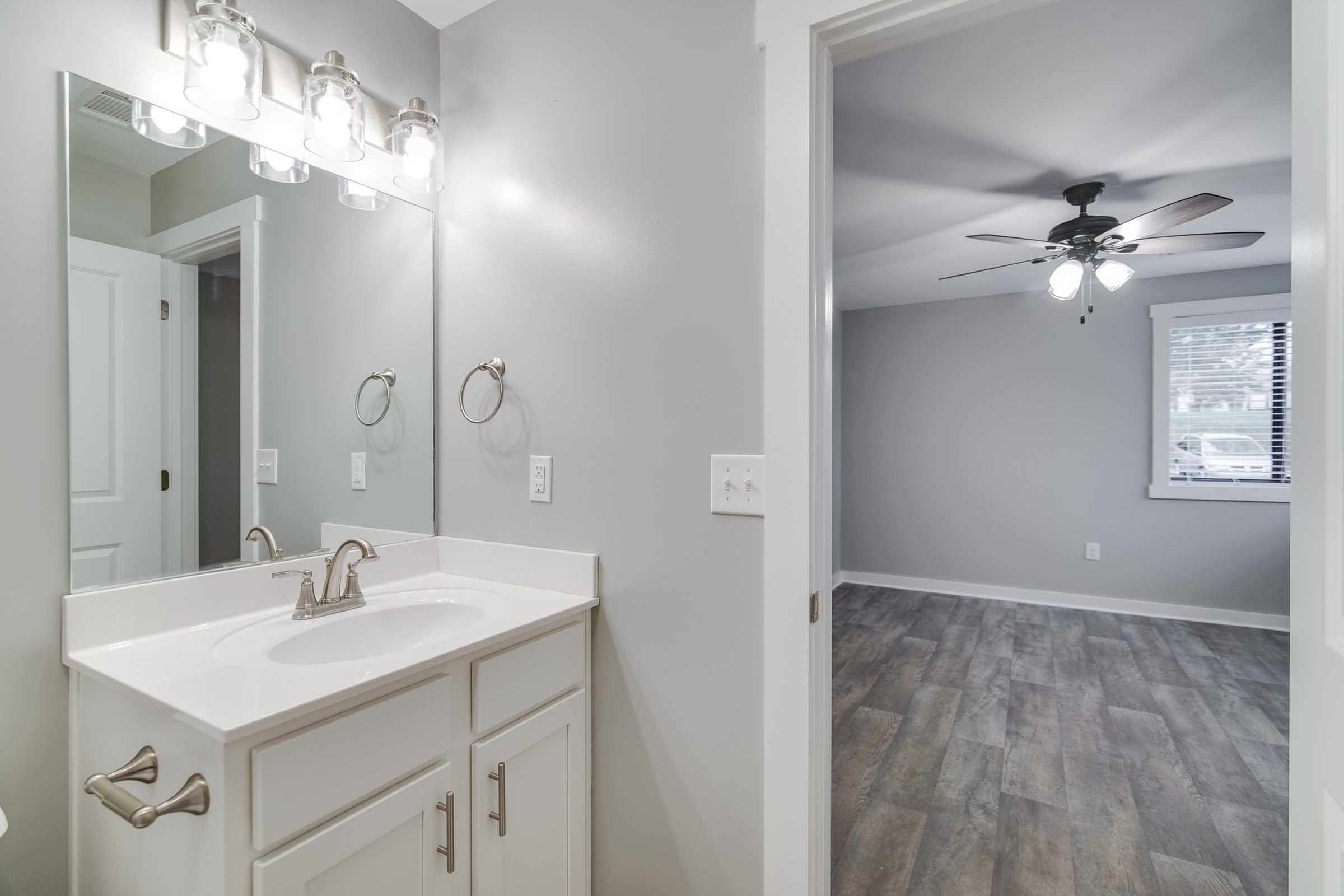 A well-lit bathroom featuring a modern vanity with a white sink and silver fixtures, a large mirror with four light bulbs above, and a door leading to a room with grey walls and a ceiling fan. The flooring is a light-colored wood laminate.