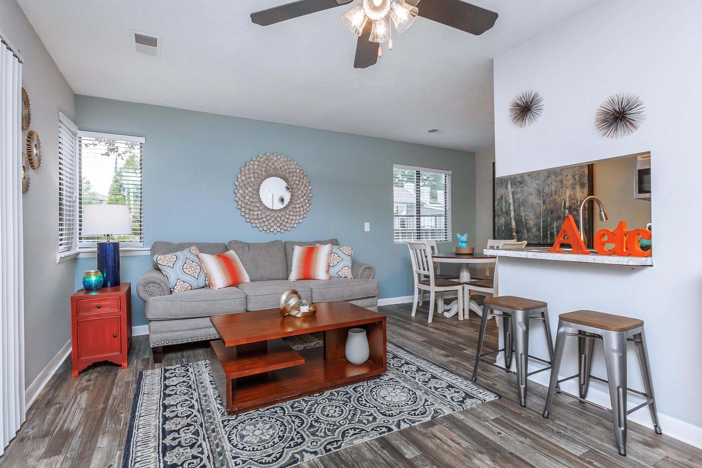 A cozy living room featuring a gray sofa with colorful throw pillows, a wooden coffee table, and a decorative area rug. There is a red accent table, stylish bar stools at a kitchen counter, and decorative wall art. Natural light fills the space through nearby windows.