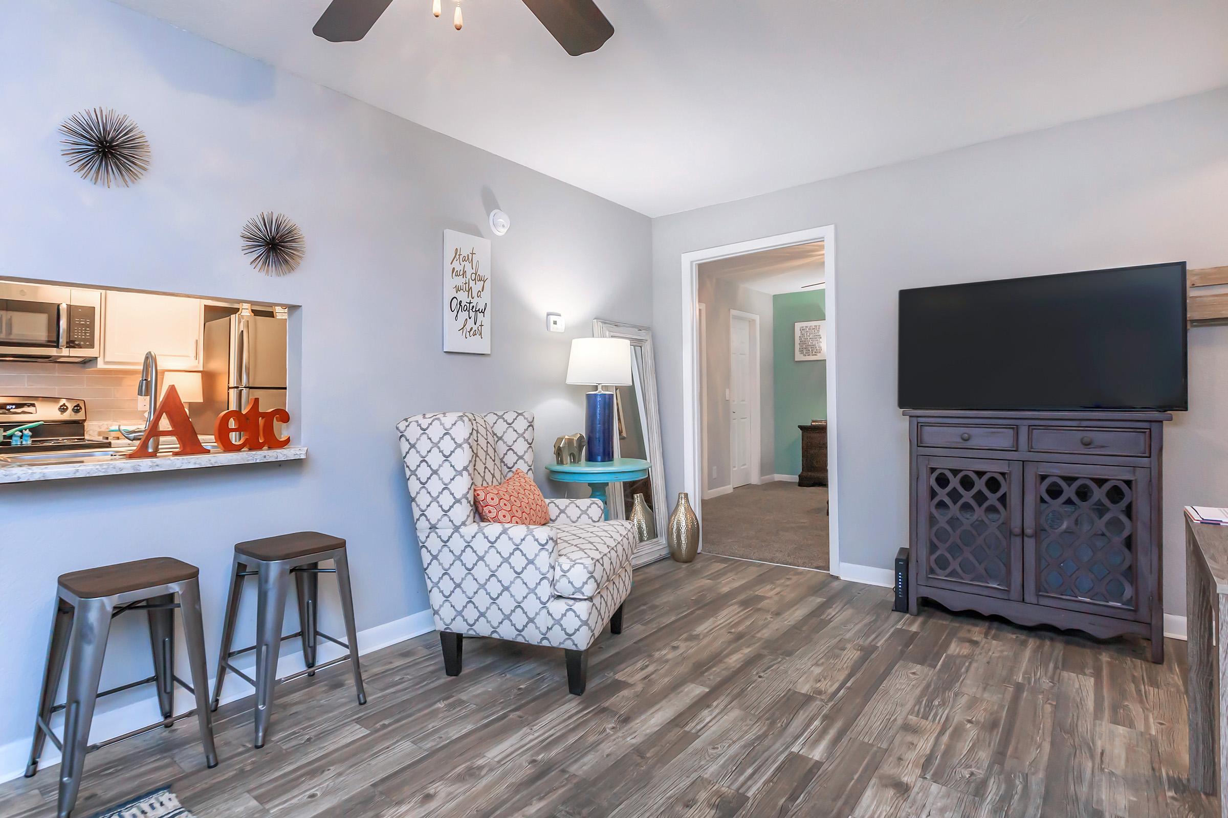 A cozy living room featuring a light gray color scheme, modern decor, and wooden flooring. It includes a patterned armchair, a small blue side table, and a TV on a stylish cabinet. The space has decorative elements on the walls and a pass-through kitchen area visible in the background, creating an inviting atmosphere.