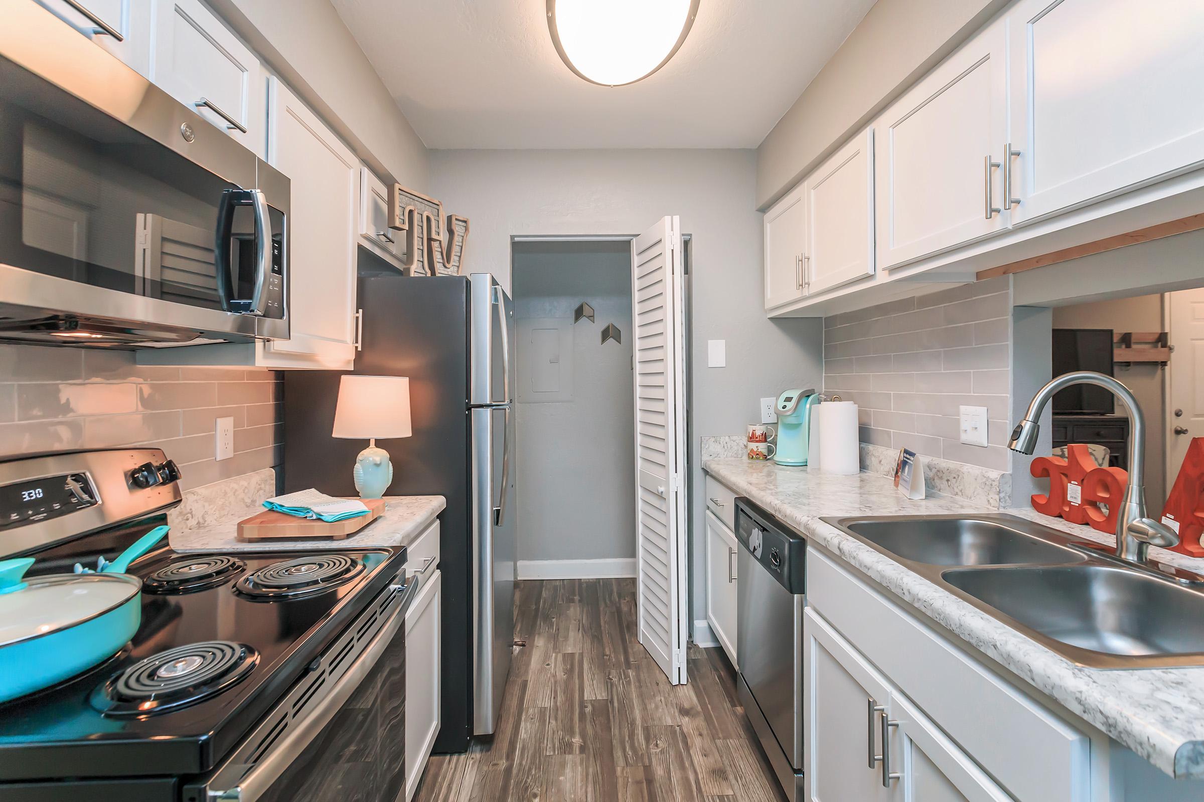 Modern kitchen with stainless steel appliances, including a stove and refrigerator. The countertops are light-colored with a patterned backsplash. There is a sink on the left and a small lamp on the counter. An open closet with folding doors is visible in the background, while decorative elements add a cozy touch.