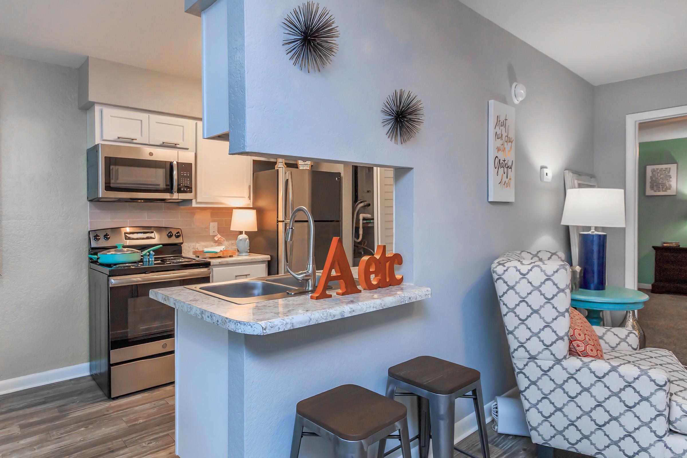 Cozy modern kitchen with stainless steel appliances, including an oven and microwave. A gray and white upholstered chair sits nearby with patterned fabric. A countertop features decorative letters and two metal stools. Soft lighting creates a welcoming atmosphere in the dining area.