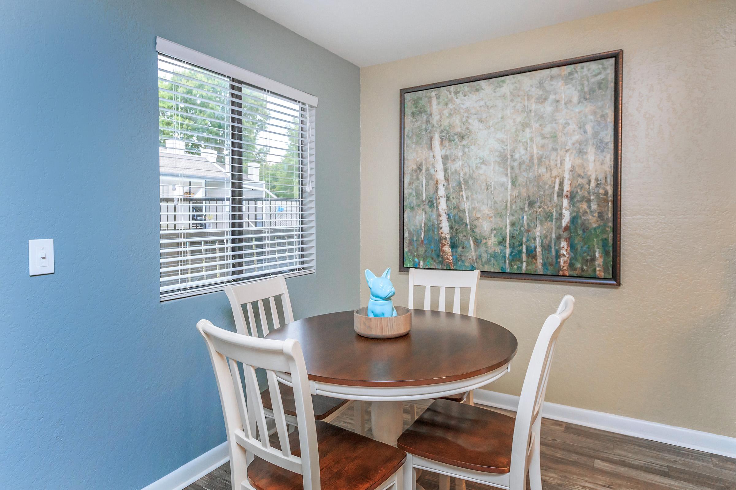 A modern dining area featuring a round wooden table with four white chairs. Centered on the table is a blue decorative figure. The wall has a large art piece depicting trees, and there are windows with white blinds allowing natural light to enter the space. The walls are painted in soft colors.