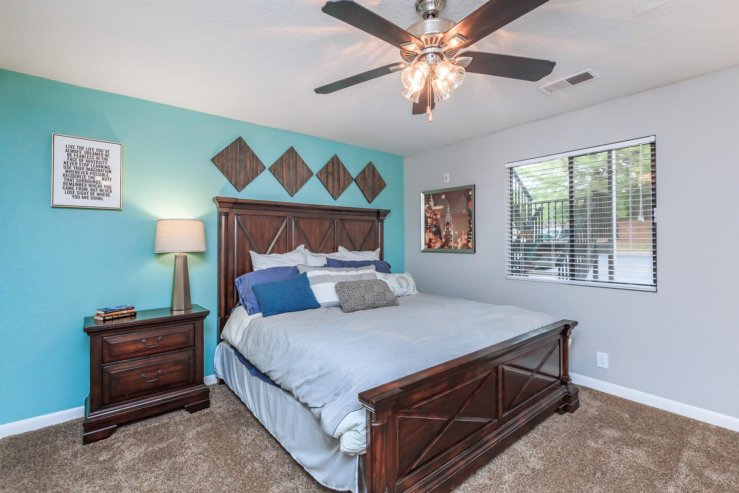 A cozy bedroom featuring a king-sized bed with blue and gray bedding, a wooden headboard, and a plush carpet. A nightstand with a lamp is beside the bed, and the wall is painted a calming blue. Decor includes geometric wood panels and framed artwork, with a window letting in natural light.
