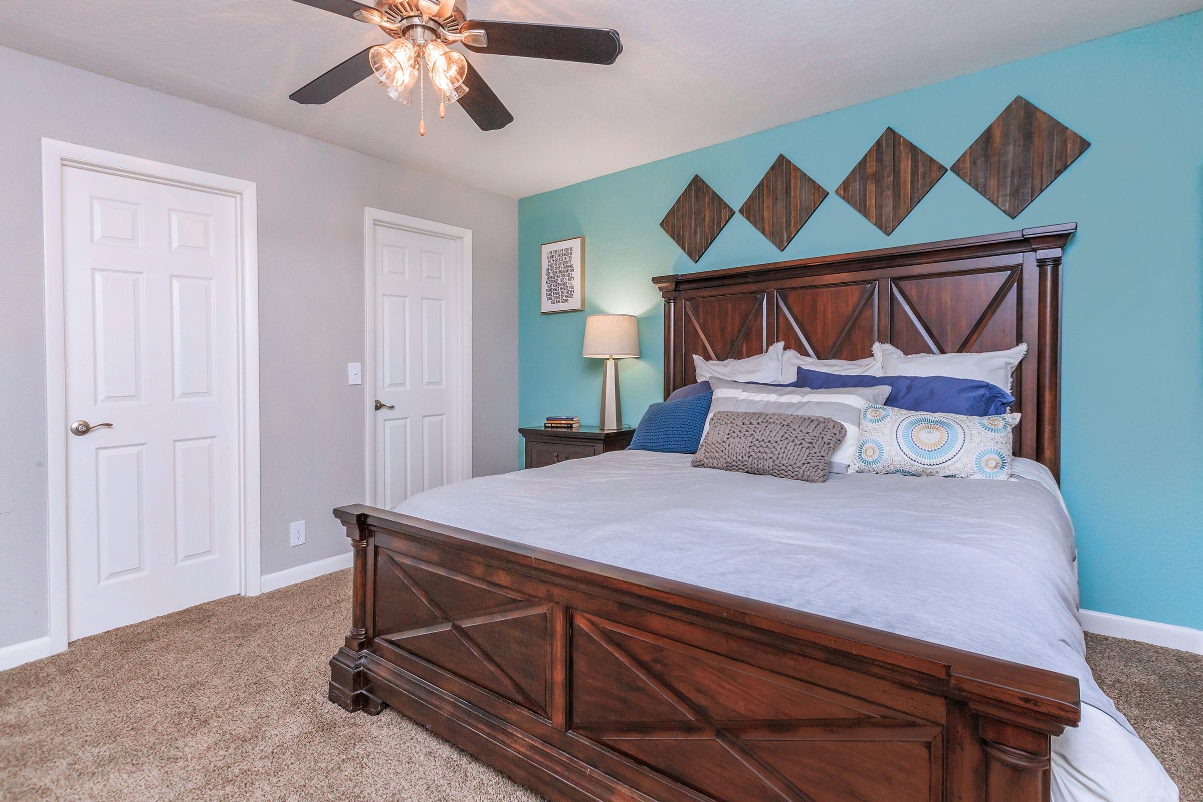 A well-decorated bedroom featuring a large wooden bed with a light gray comforter, decorative pillows, and a nightstand. The walls are painted in gray and teal, and there are geometric wall decorations above the bed. A ceiling fan with lights hangs above, and two white doors are visible on the left.