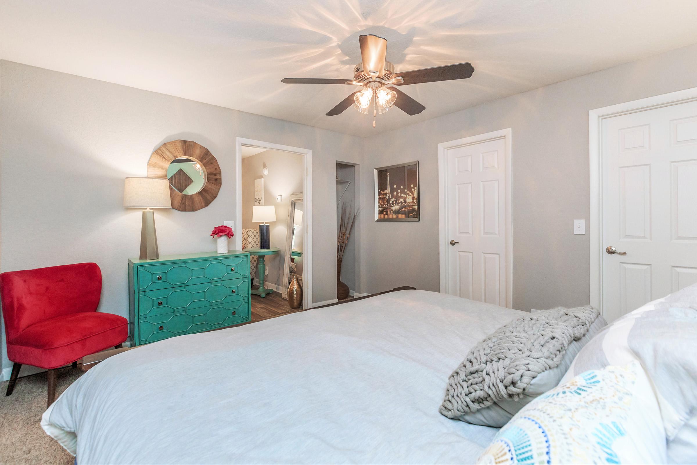 A cozy bedroom featuring a light blue bedspread, a decorative ceiling fan, and a teal dresser. A red accent chair adds a pop of color. A lamp is on a bedside table, and there are two closed doors leading to other rooms. Soft gray walls create a warm ambiance.