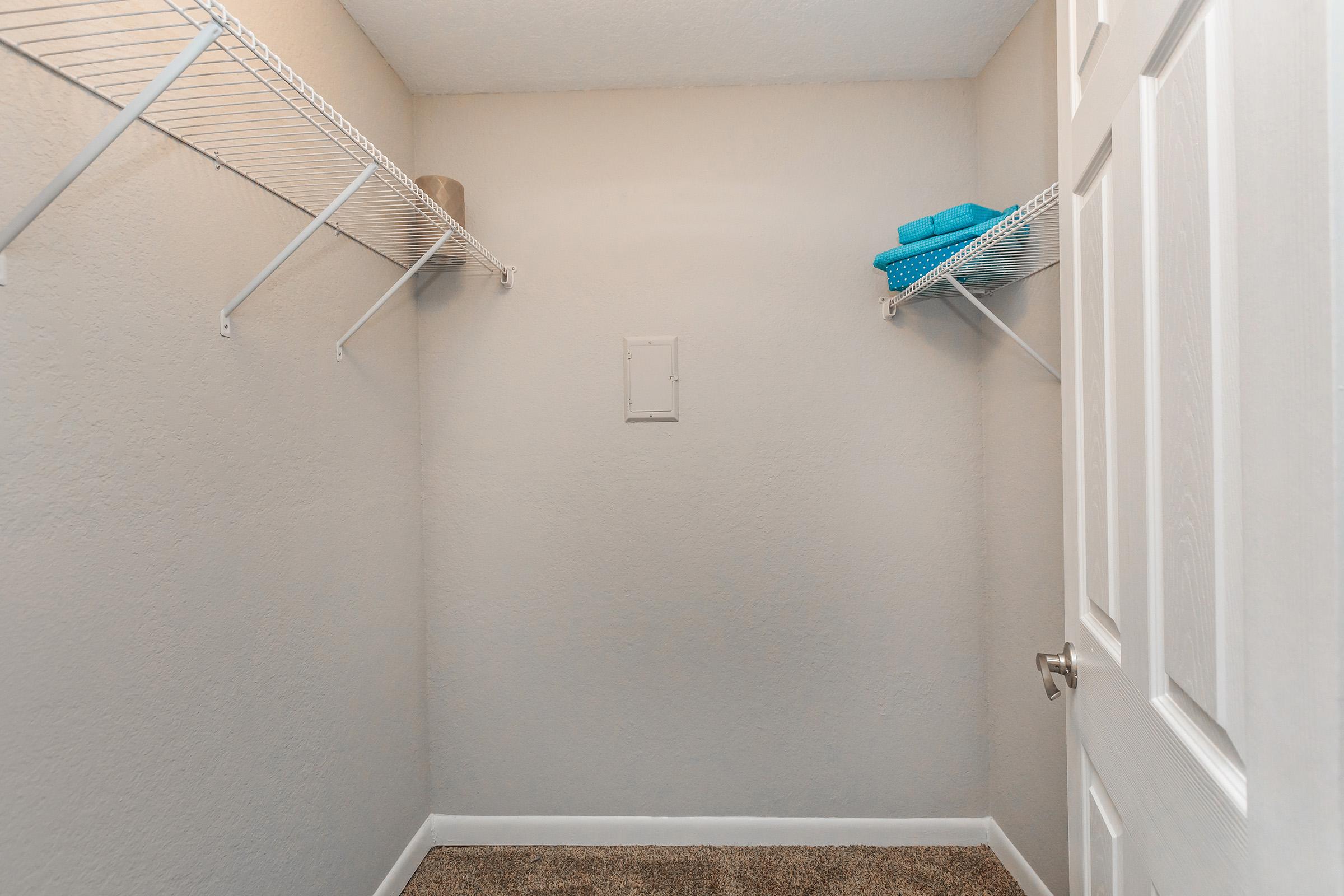 A small closet with light gray walls and a carpeted floor. It features two wire shelves on the left side, one holding a roll of paper and the other with neatly folded blue towels. A white door partially closed is visible, and there is no furniture or additional items in the space.