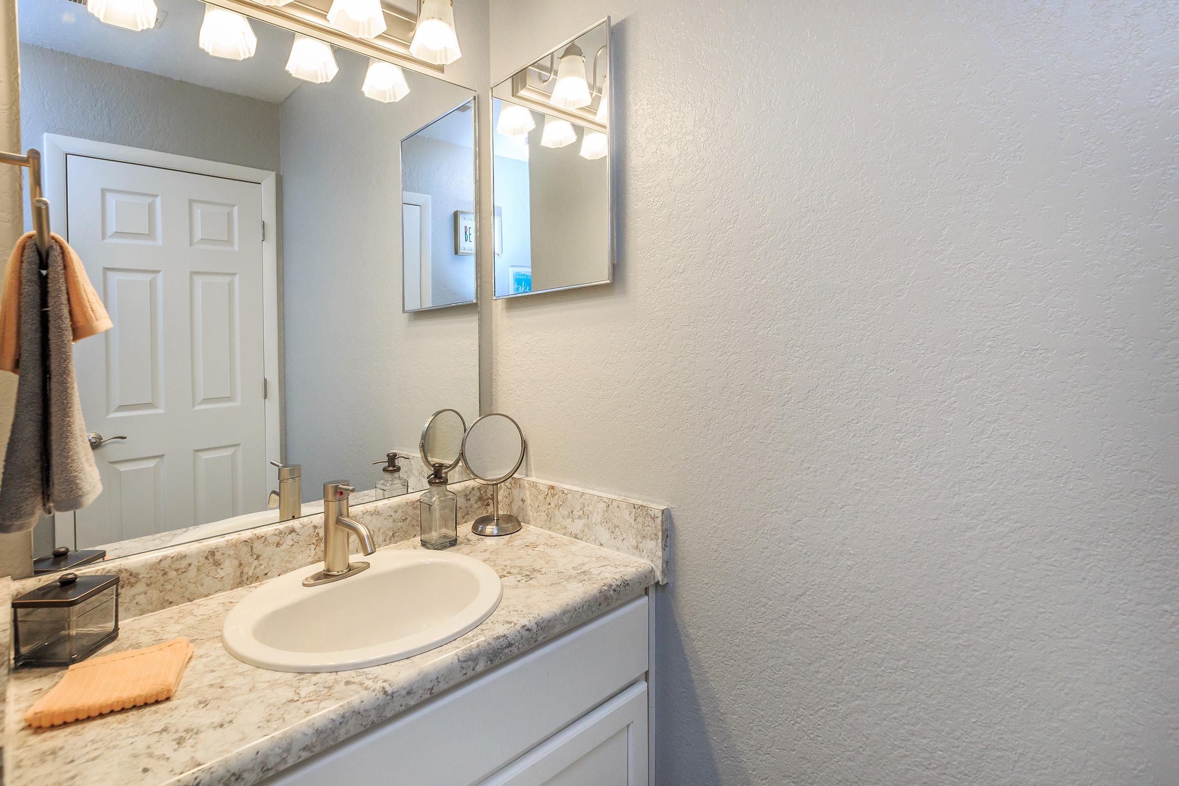 A clean and modern bathroom featuring a white sink with a circular mirror, a countertop with grey-patterned surface, and a set of light fixtures above. Soft towels hang nearby, and there is a small clear container on the counter, enhancing the organized and minimalist look of the space.
