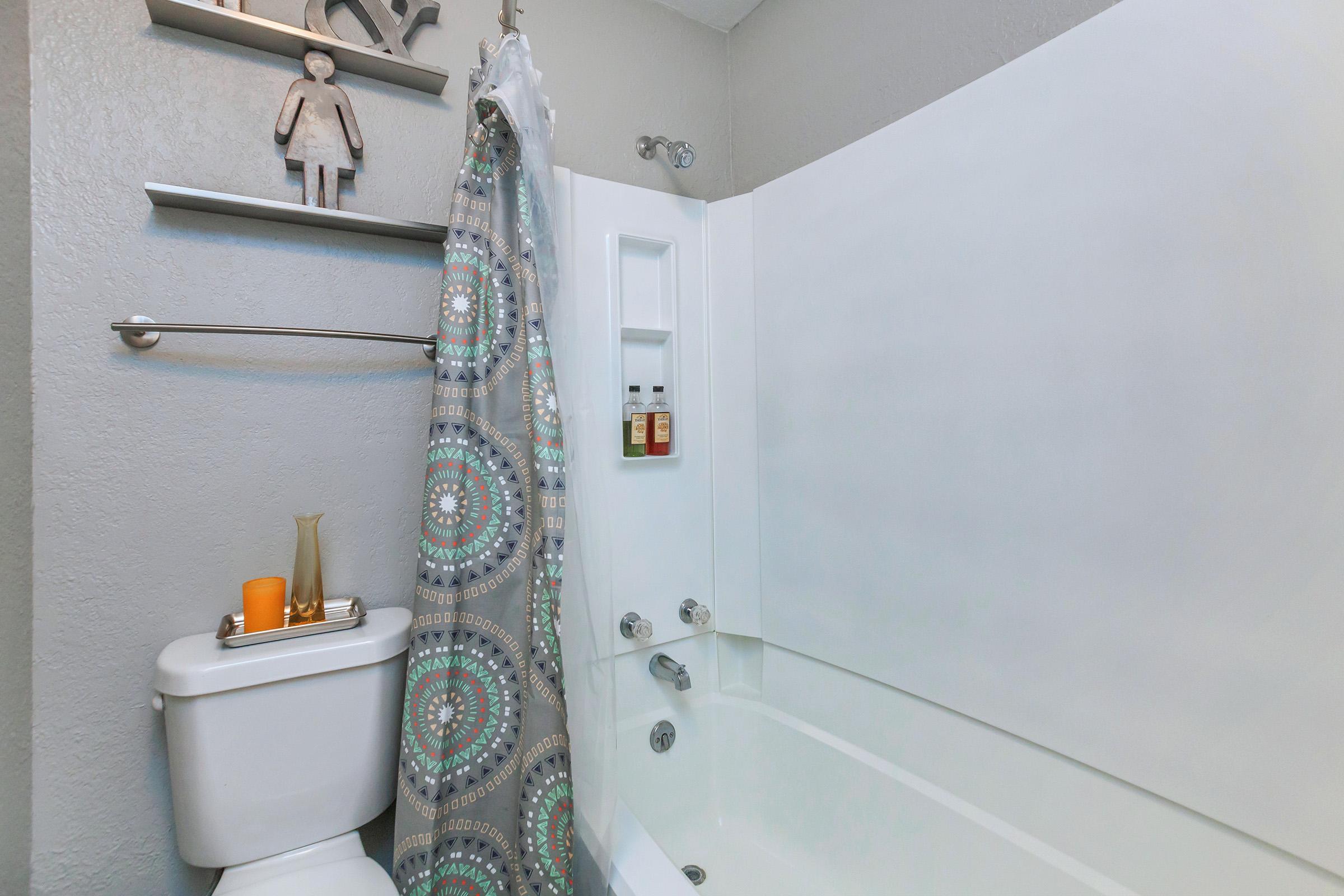 A modern bathroom featuring a bathtub with a shower curtain displaying a colorful pattern. There's a shelf with decorative items, and a toilet is positioned nearby. The wall is painted in a light gray color, creating a clean and contemporary aesthetic.