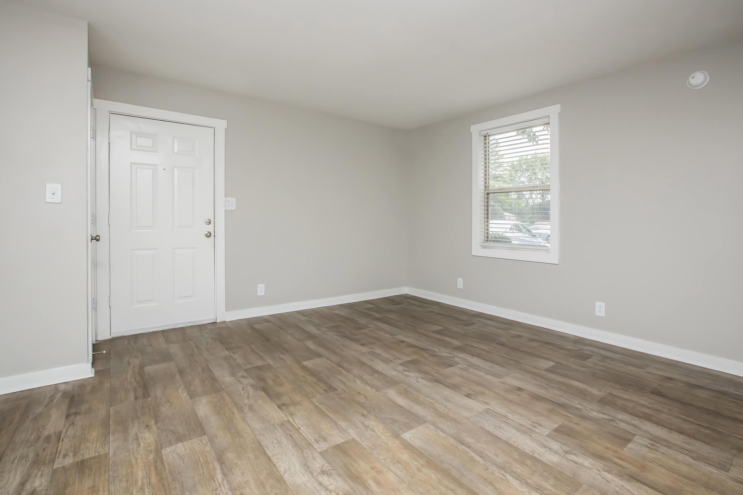 A spacious, empty room with light gray walls, a white door leading outside, and a window with blinds. The floor features wood-like laminate, giving a warm appearance. There's no furniture, creating a clean, minimalist look, ideal for potential decorating.