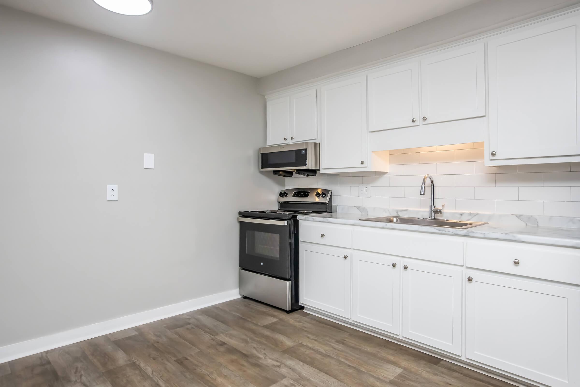 Modern kitchen featuring white cabinetry, stainless steel appliances including an oven and microwave, a sink with a faucet, and a marble countertop. The walls are painted a light gray, and the floor has wooden laminate planks, creating a clean and contemporary look.