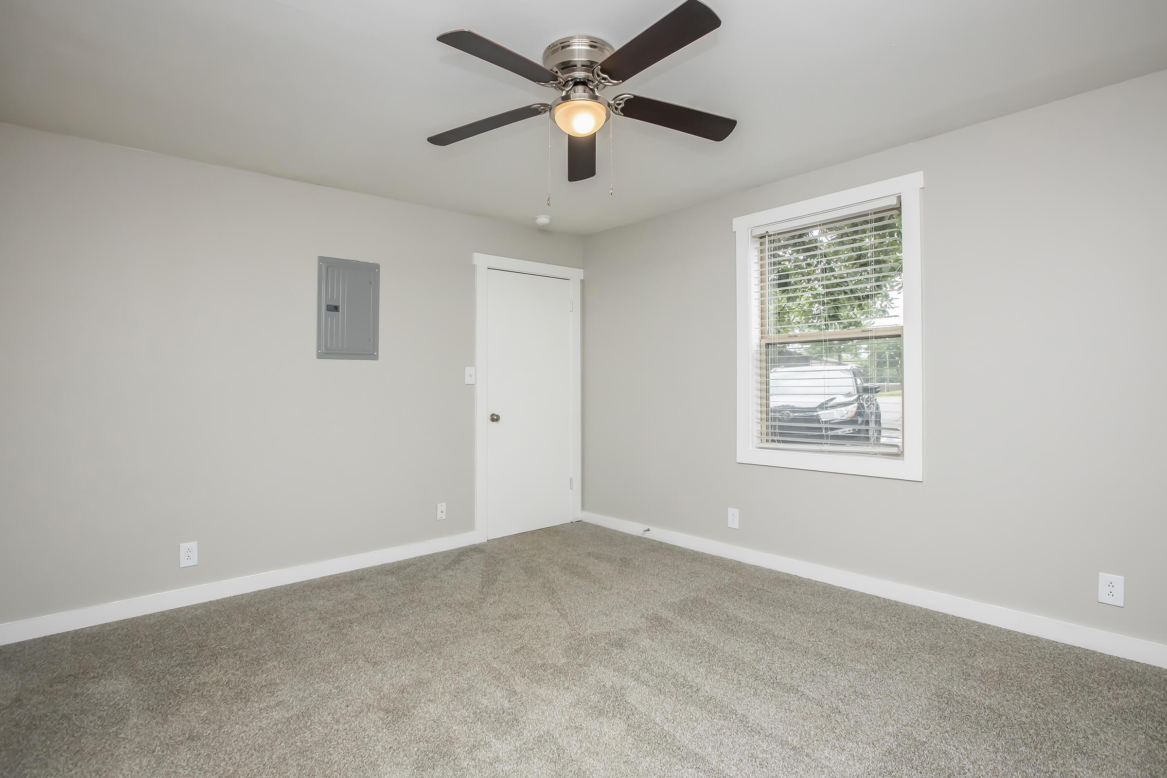 Empty room with gray walls and a light gray carpet. It features a ceiling fan with light, a small electrical panel on the wall, and a window with white blinds. A white door is visible, leading to another space. The room has a quiet and spacious feel.