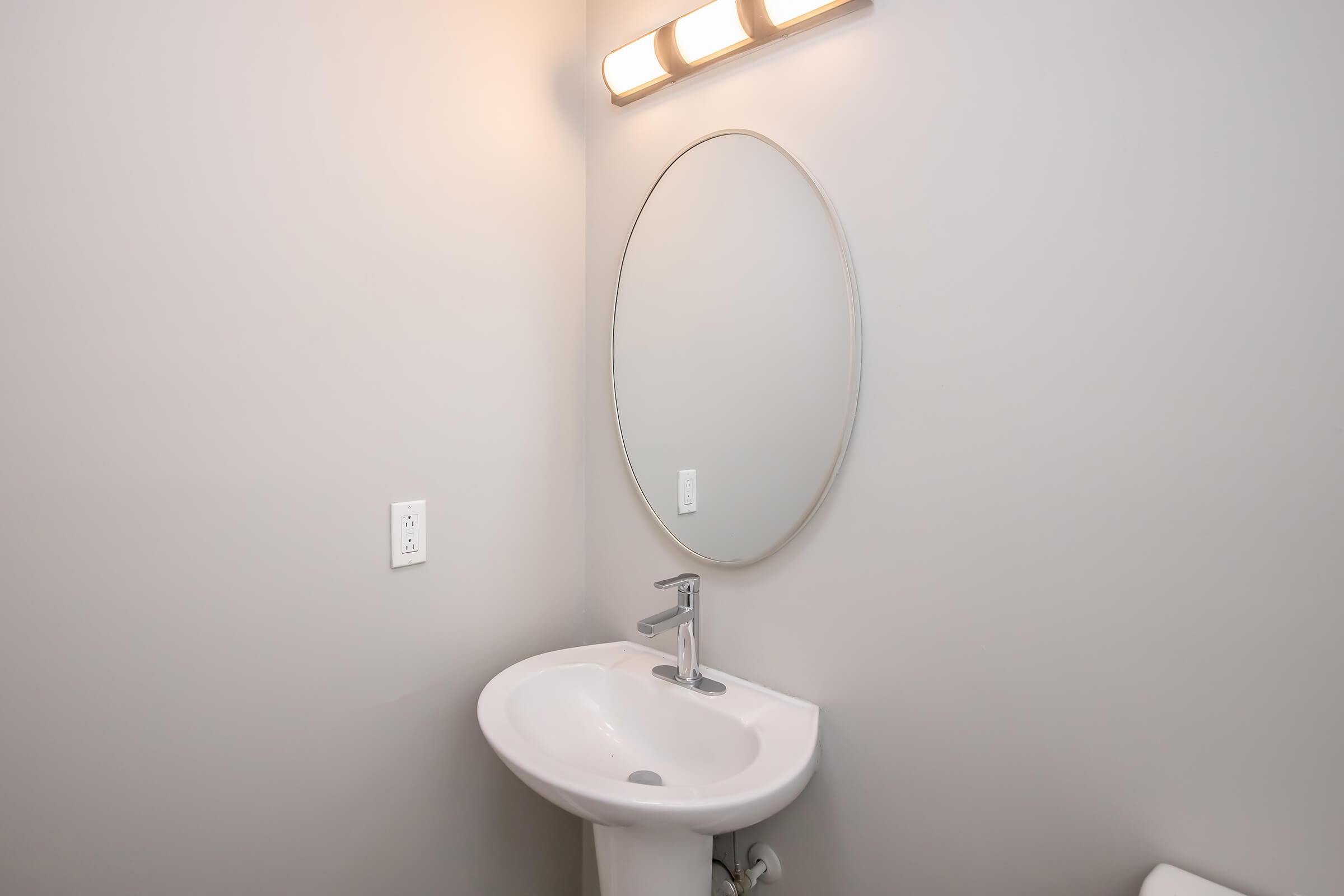 A modern bathroom with light gray walls, featuring a round mirror above a white pedestal sink. A sleek faucet is visible, and there's a wall-mounted light fixture providing soft illumination. The overall design is minimalistic and contemporary.