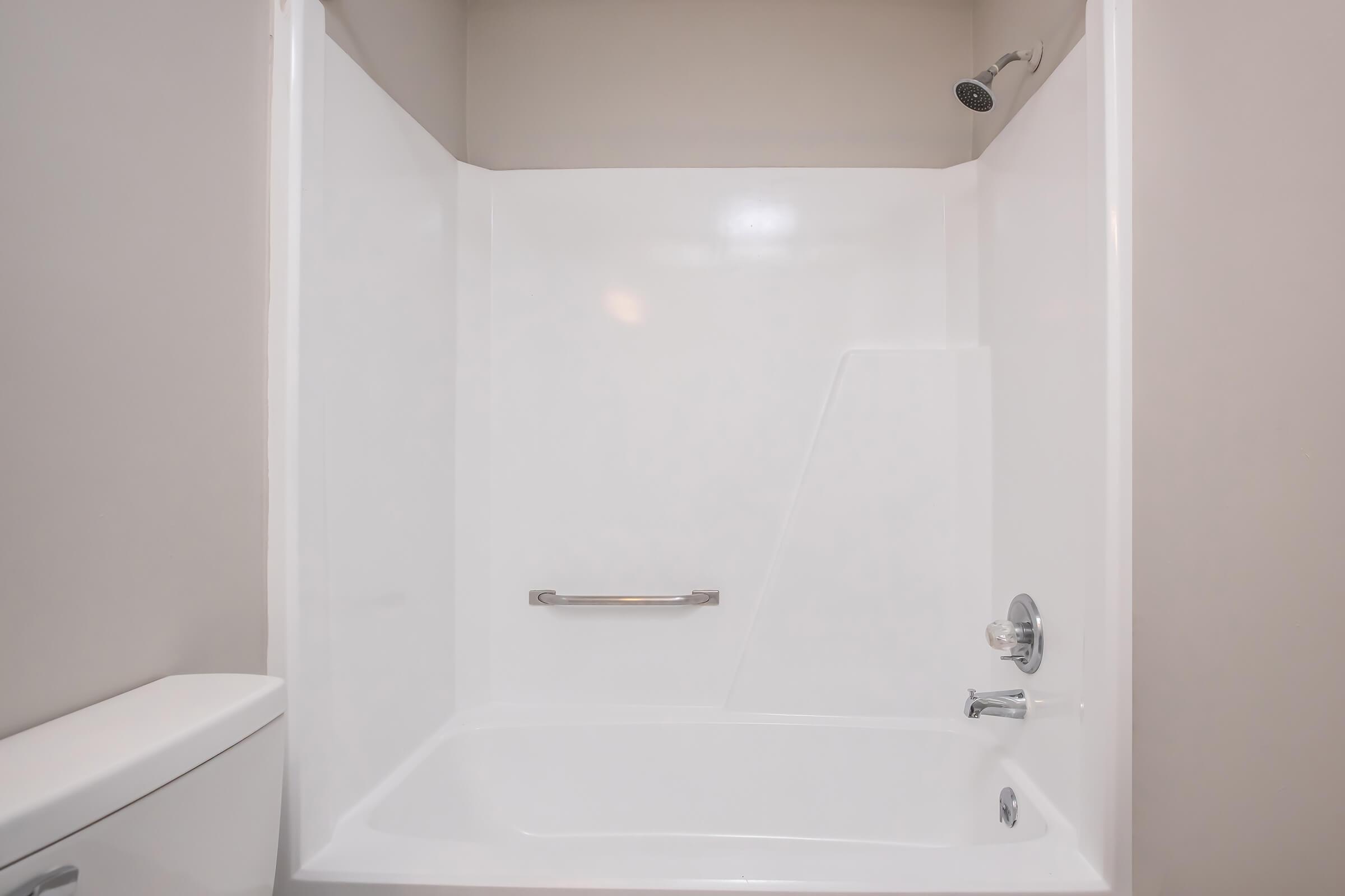 A clean, empty bathroom shower space with a white tub and shower surround. The shower features a wall-mounted faucet and a handheld showerhead. A grab bar is installed for safety, and the wall is painted a neutral color, creating a bright and airy atmosphere.