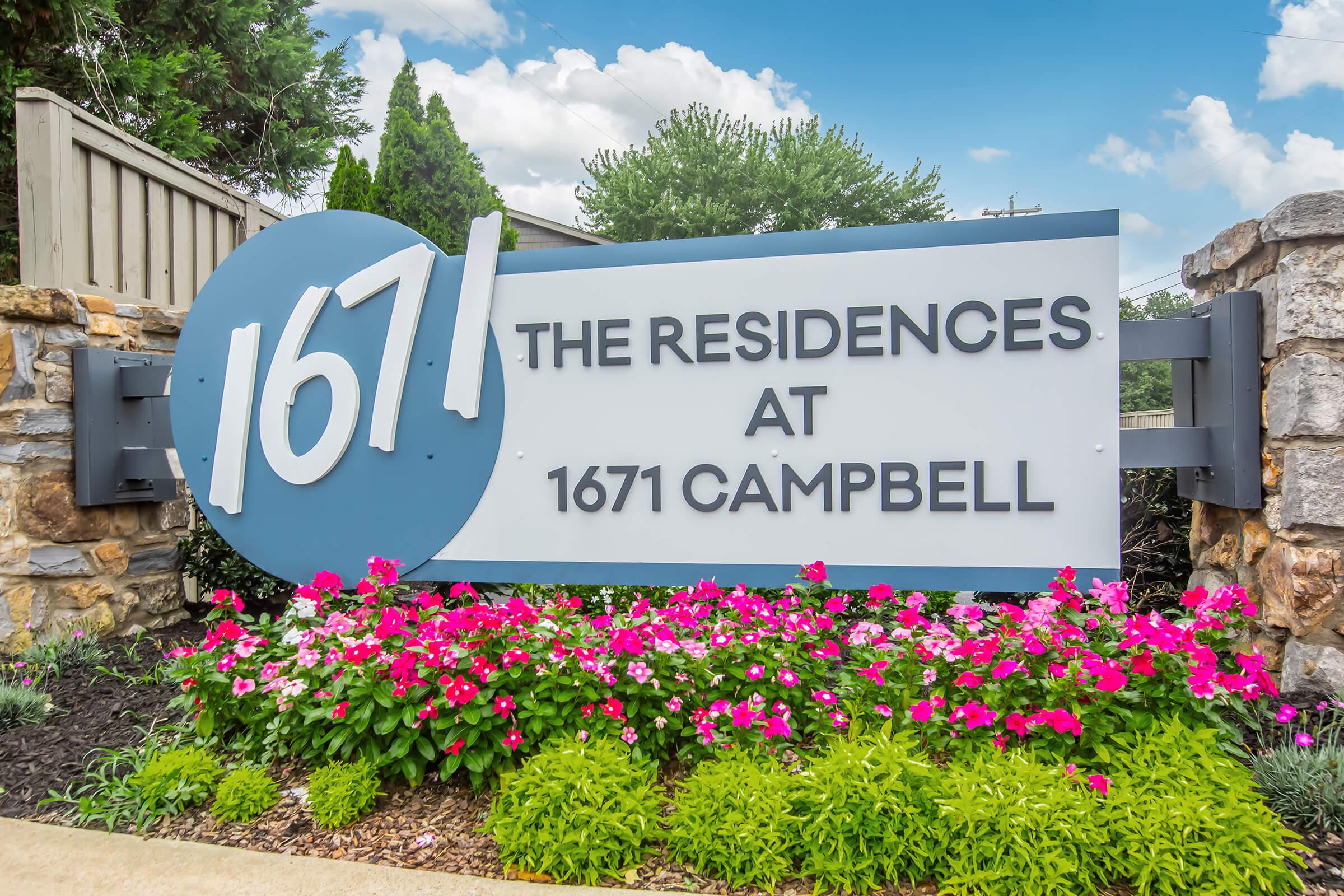 Sign for "The Residences at 1671 Campbell," featuring a large circular blue graphic with the number 1671, surrounded by vibrant pink and purple flowers. The background includes green shrubbery and a clear blue sky.